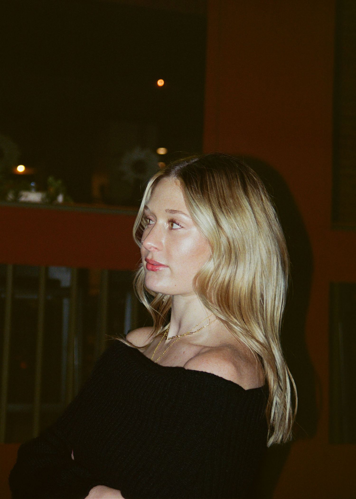 Blonde woman in black off-shoulder top gazing away in warm ambient evening light.