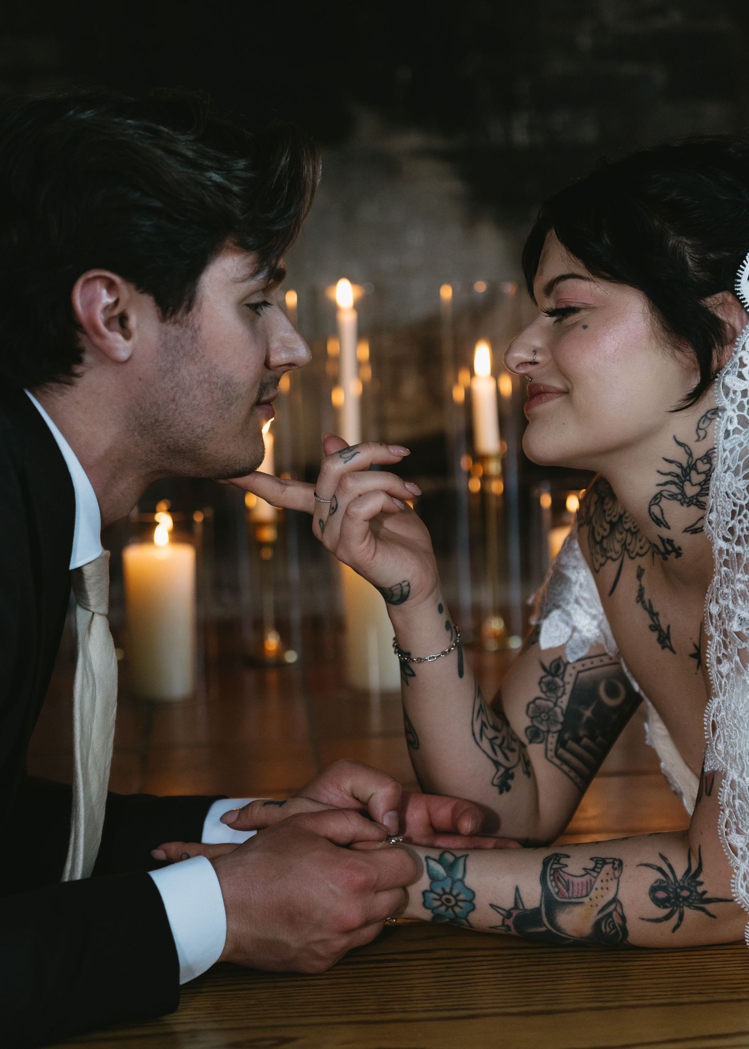Tattooed bride and groom share an intimate moment at candlelit table, holding hands and gazing at each other.