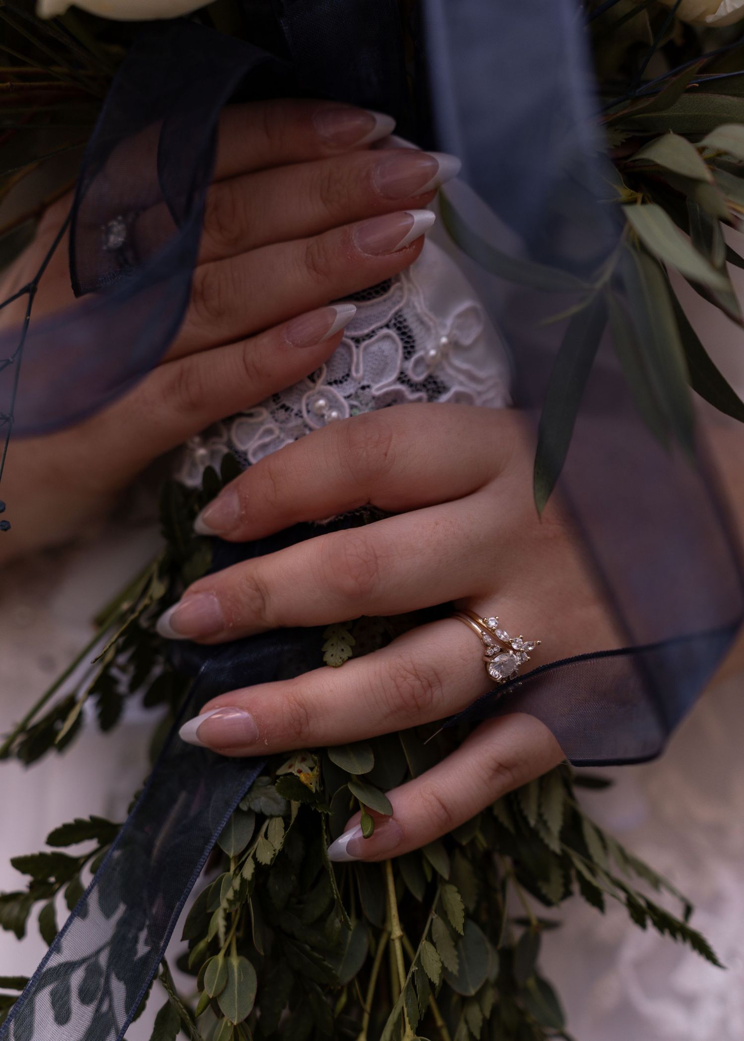 Close-up of bride holding bouquet with greenery, wearing gold diamond engagement ring and nude nails.