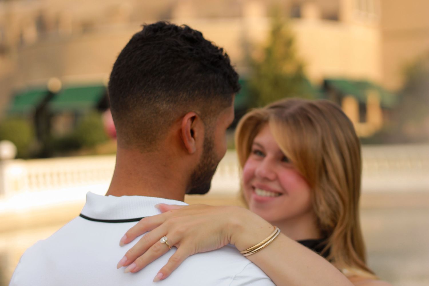 Smiling blonde woman hugs her partner outdoors, showing off her engagement ring.