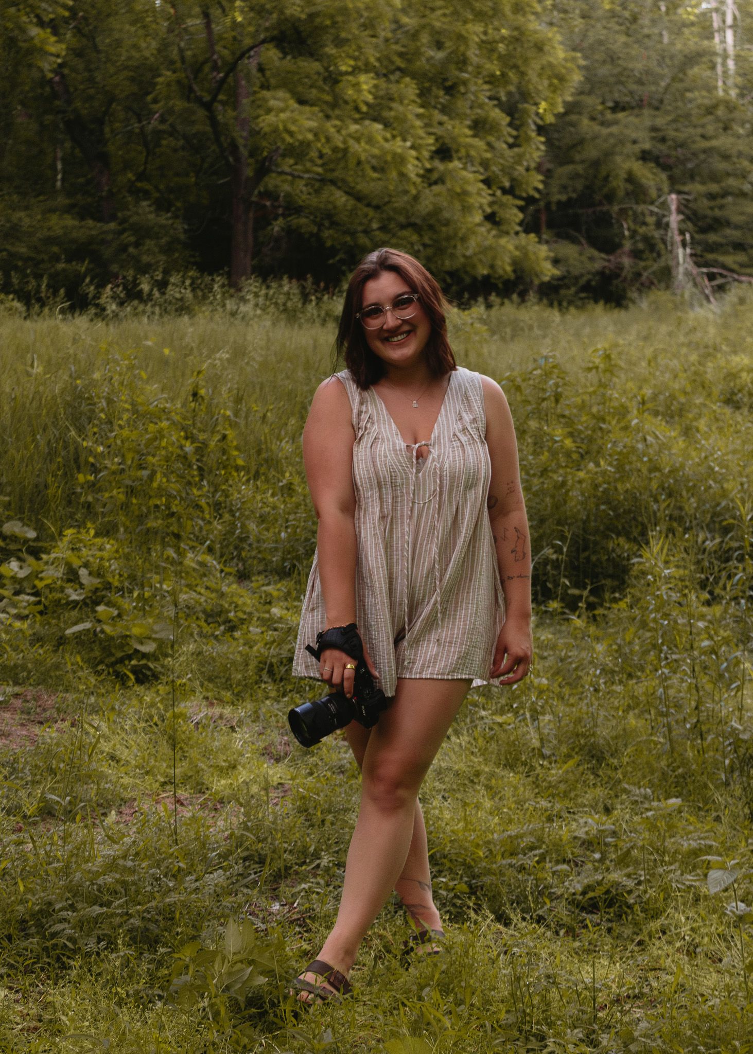 Young woman in striped romper holding a camera, smiling outdoors in a lush green forest meadow.