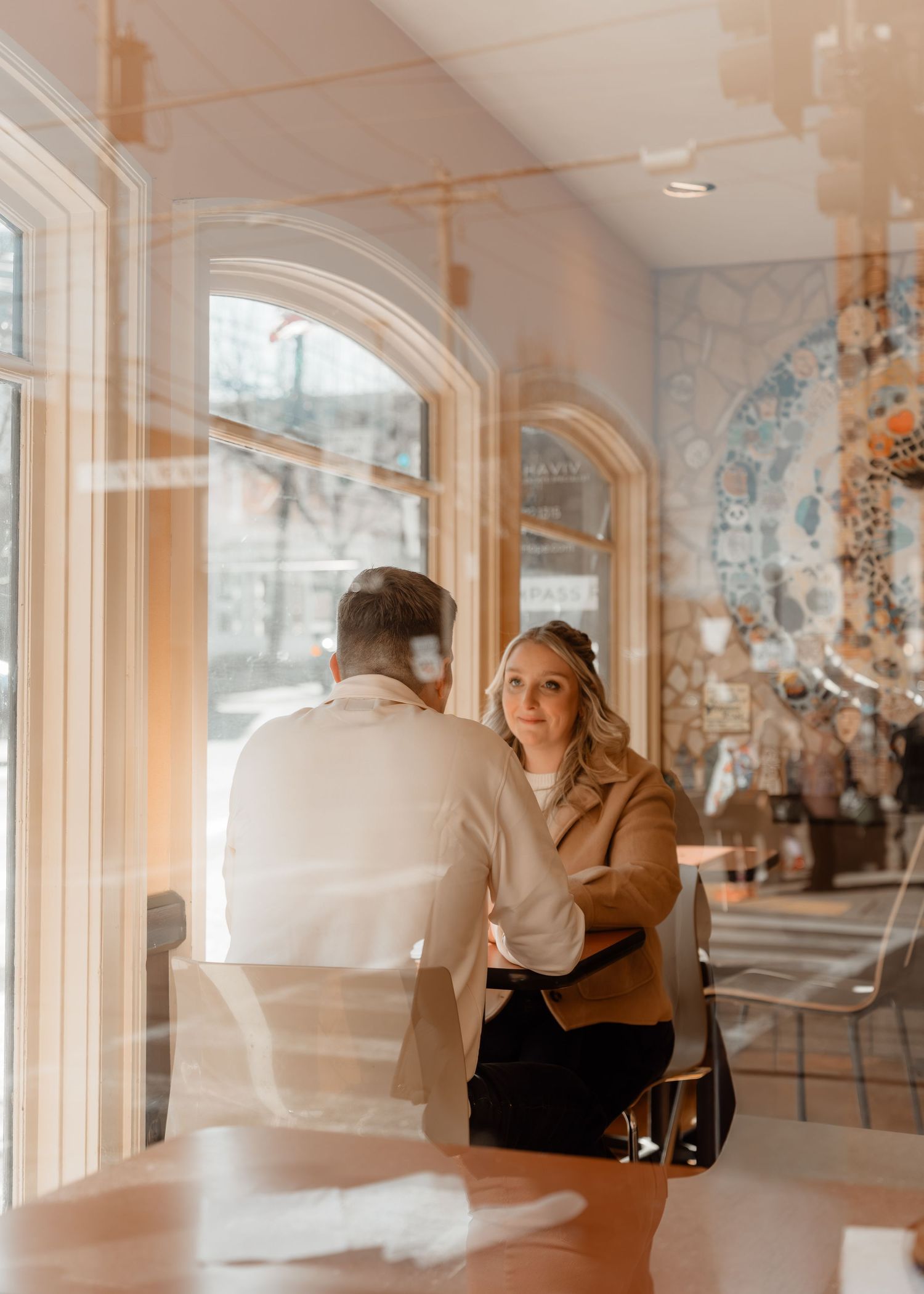 Couple sharing an intimate moment at a cafe table, photographed through a window with warm light reflections.
