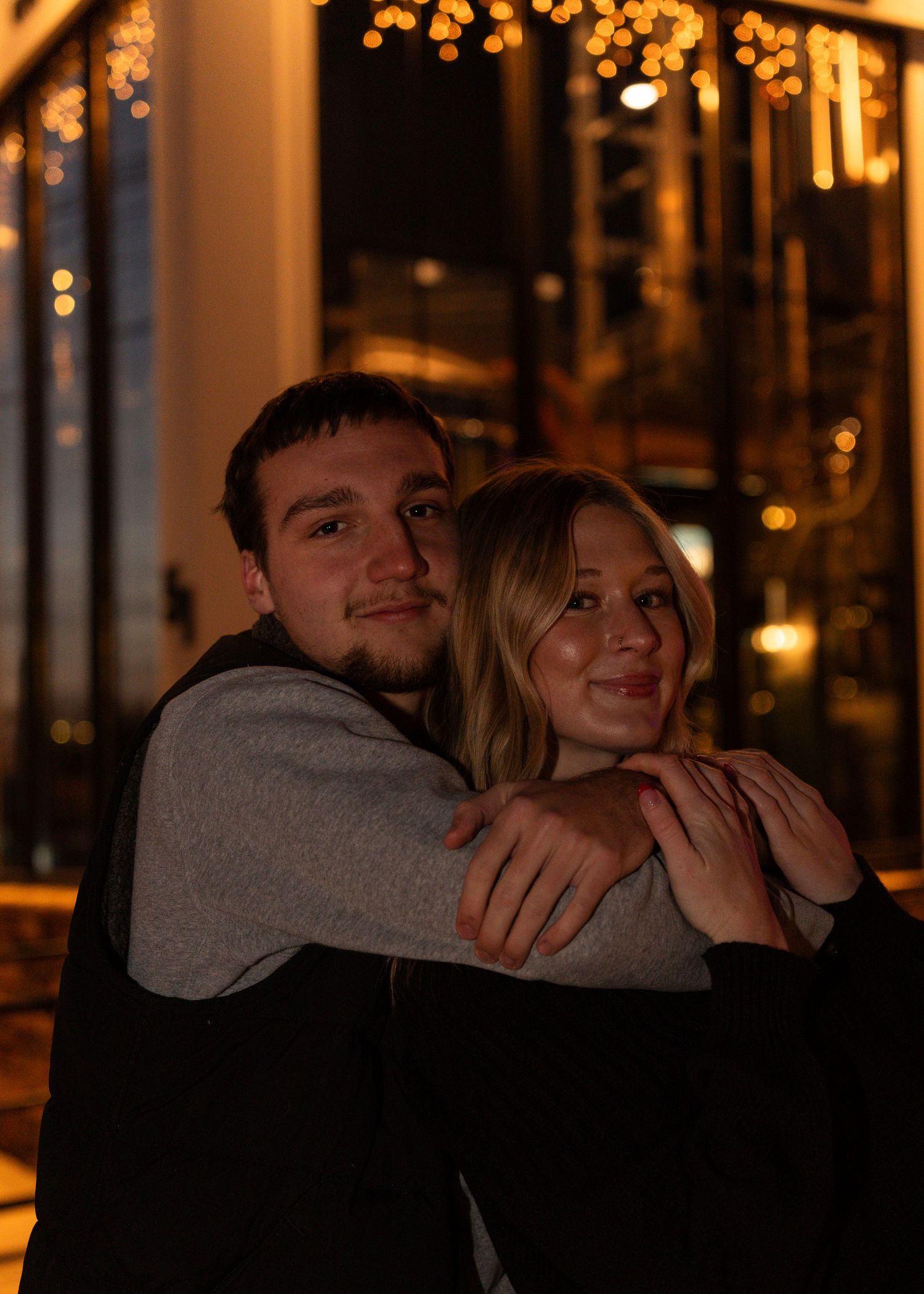 Young couple embracing and smiling outdoors at night with warm bokeh lights in the background.
