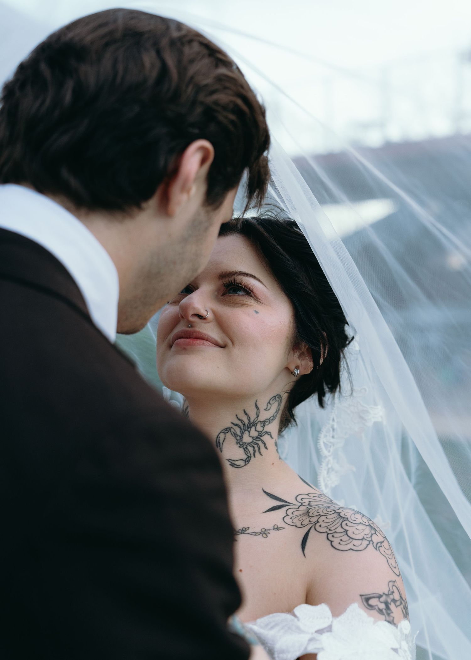 Tattooed bride with scorpion neck tattoo gazes up at groom under flowing white wedding veil outdoors.
