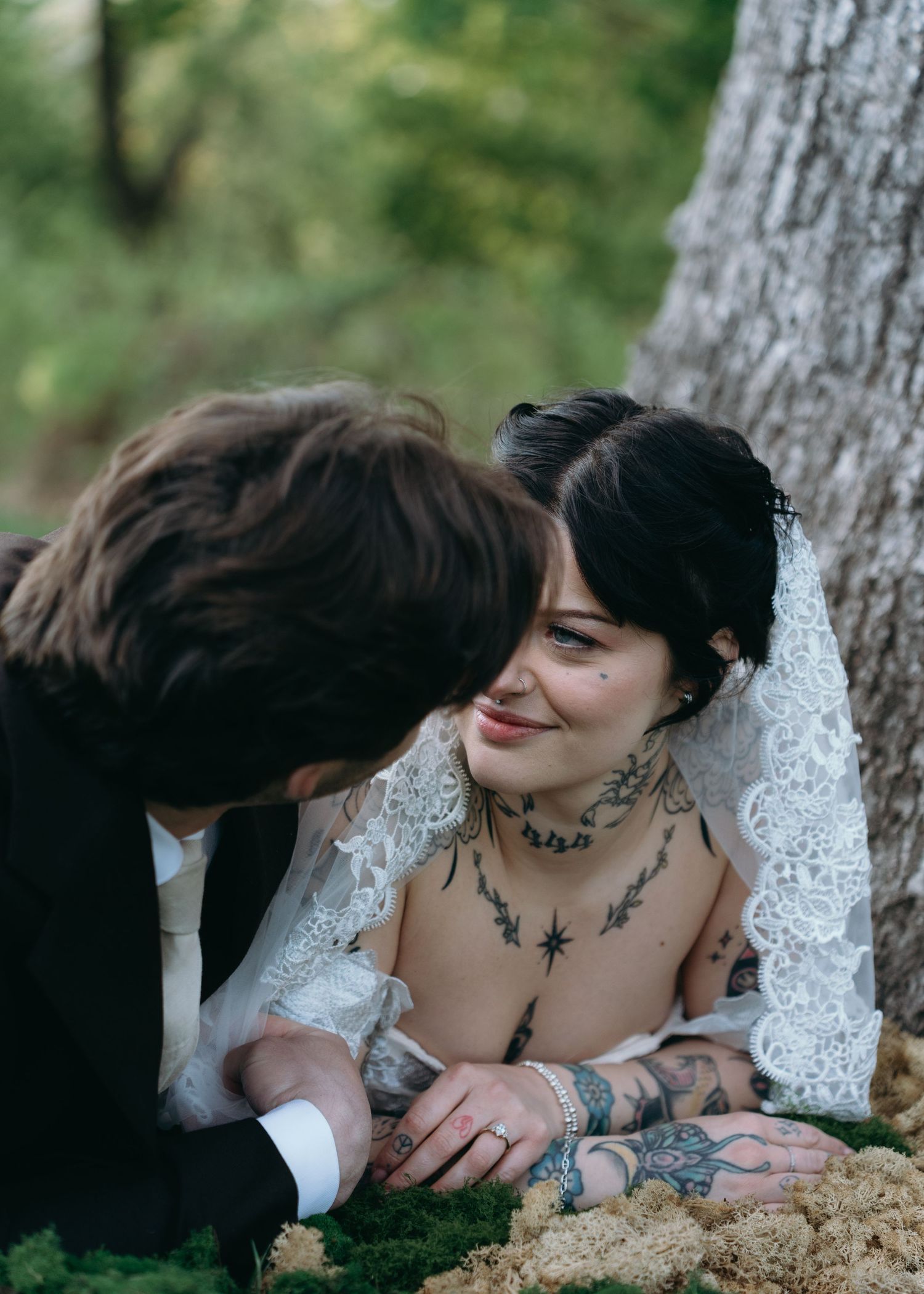 Tattooed bride smiles as groom leans in to kiss her cheek while sitting against a tree in the woods.