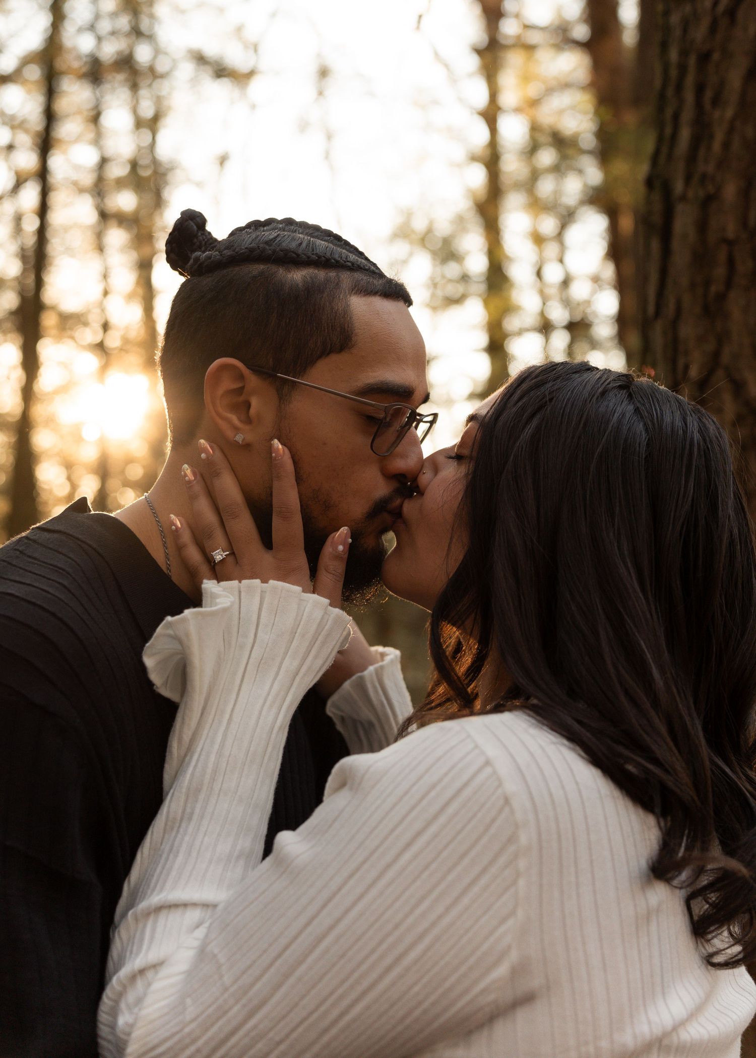 Couple sharing a romantic kiss at golden hour in a sunlit forest, woman wearing engagement ring.