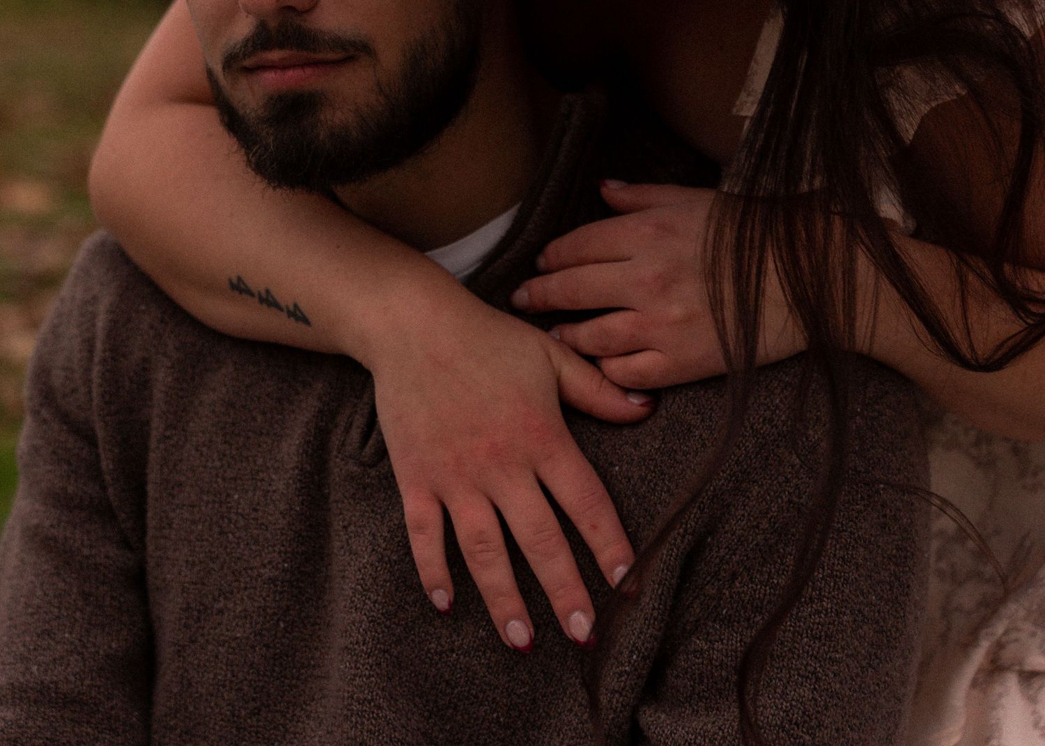 Close-up of a couple embracing outdoors, woman's hand resting on man's shoulder with small tattoo.