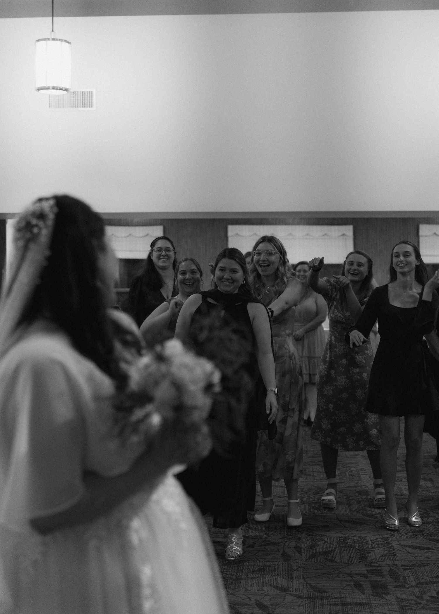 Black and white photo of bride preparing to toss bouquet to excited group of women at reception.