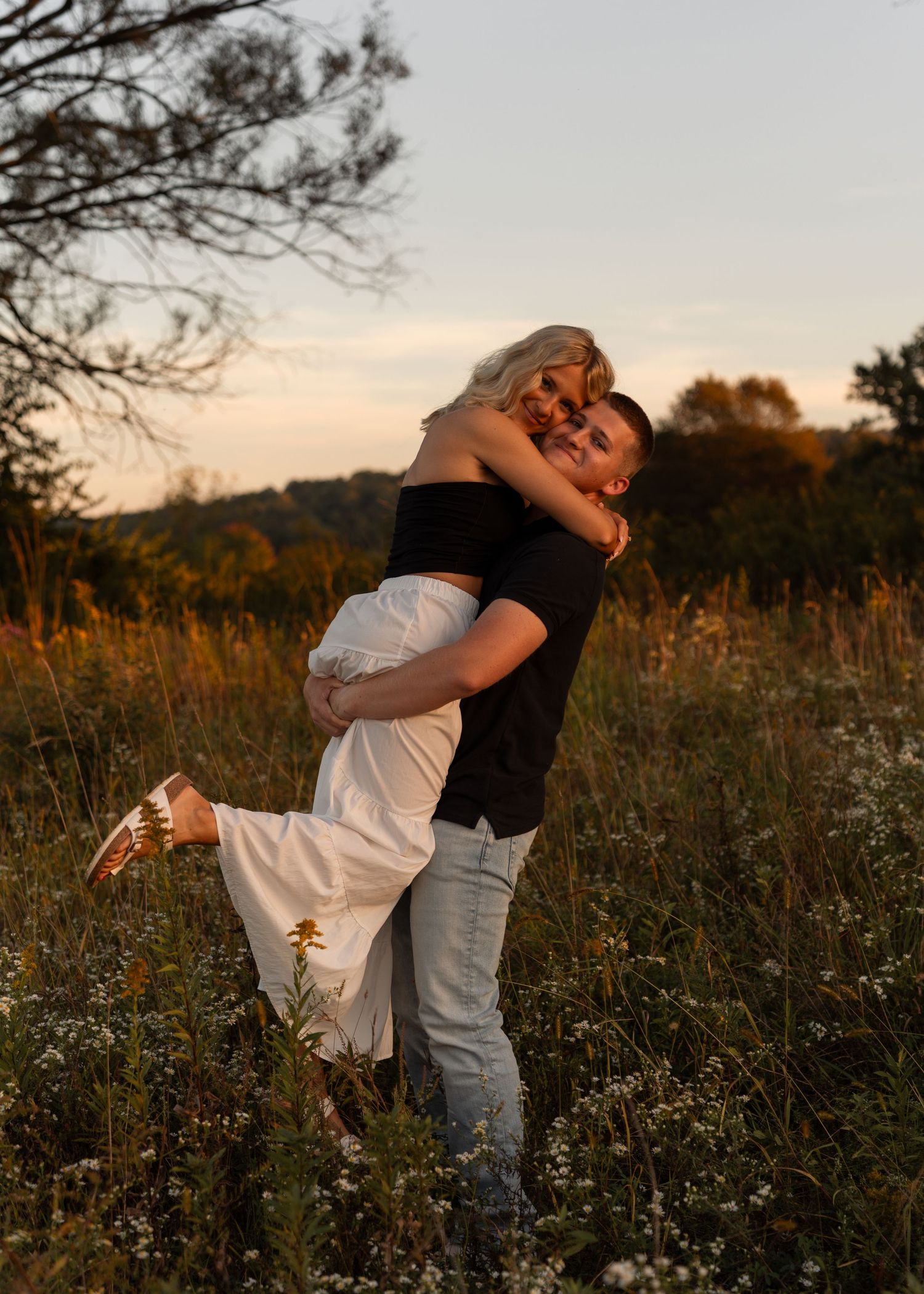 A couple embraces joyfully in a golden wildflower field at sunset, man lifting woman in white skirt.