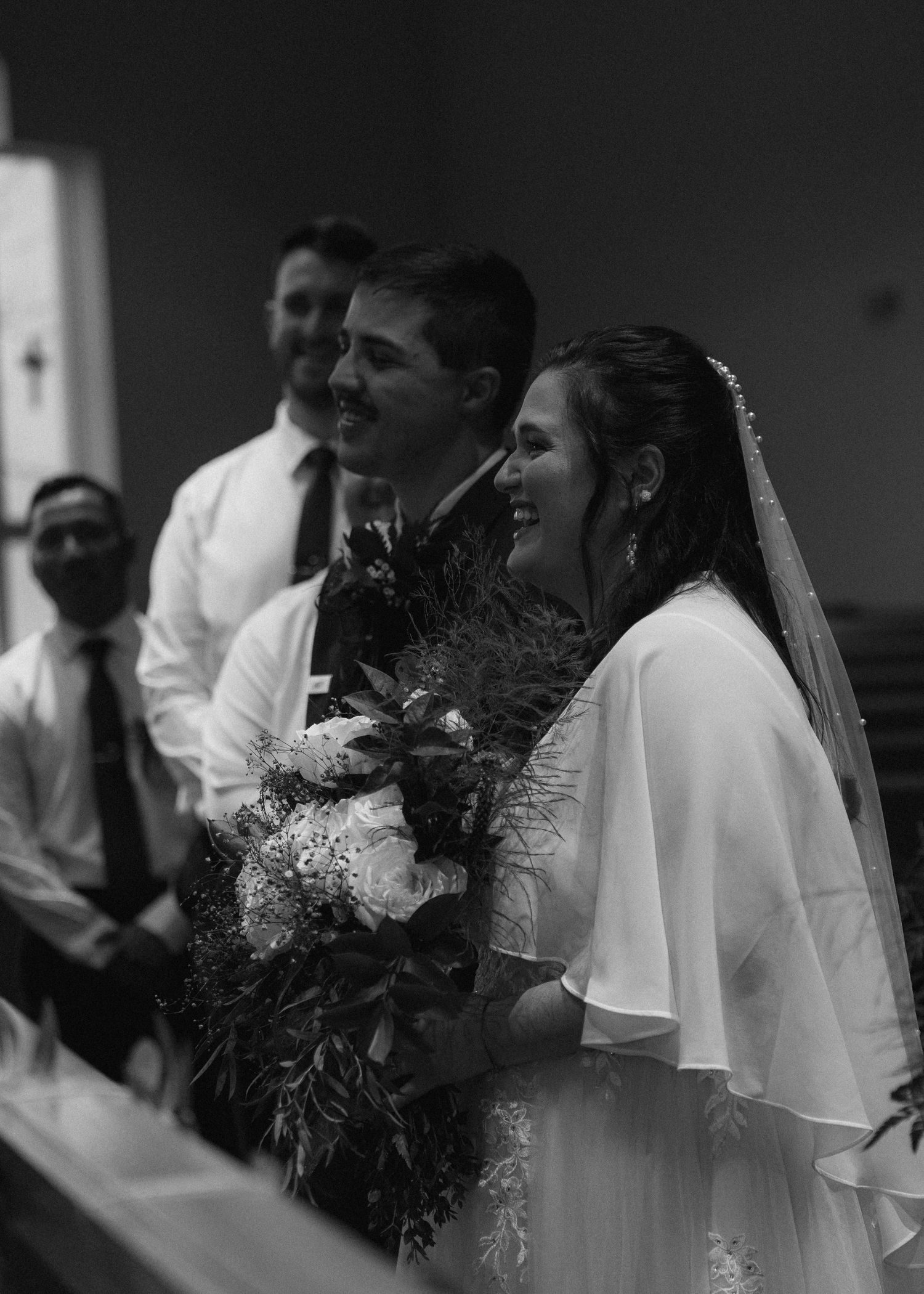 Black and white photo of smiling bride and groom holding bouquet during wedding ceremony at altar.