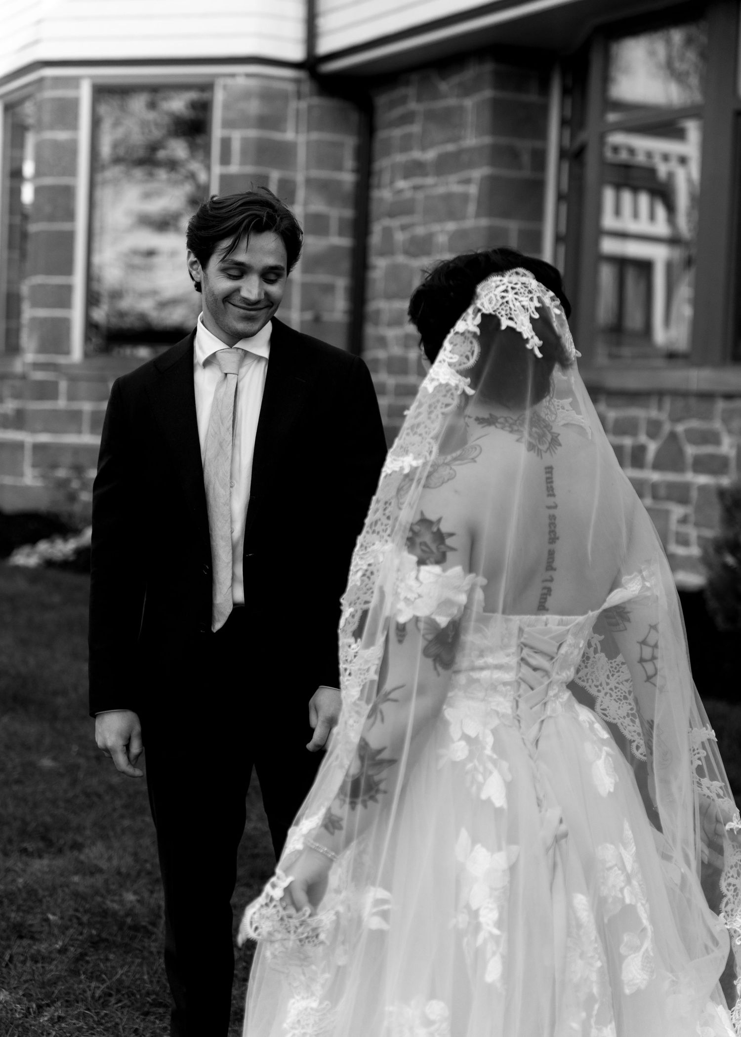 Black and white photo of smiling groom in suit walking beside bride in lace veil outside stone building.