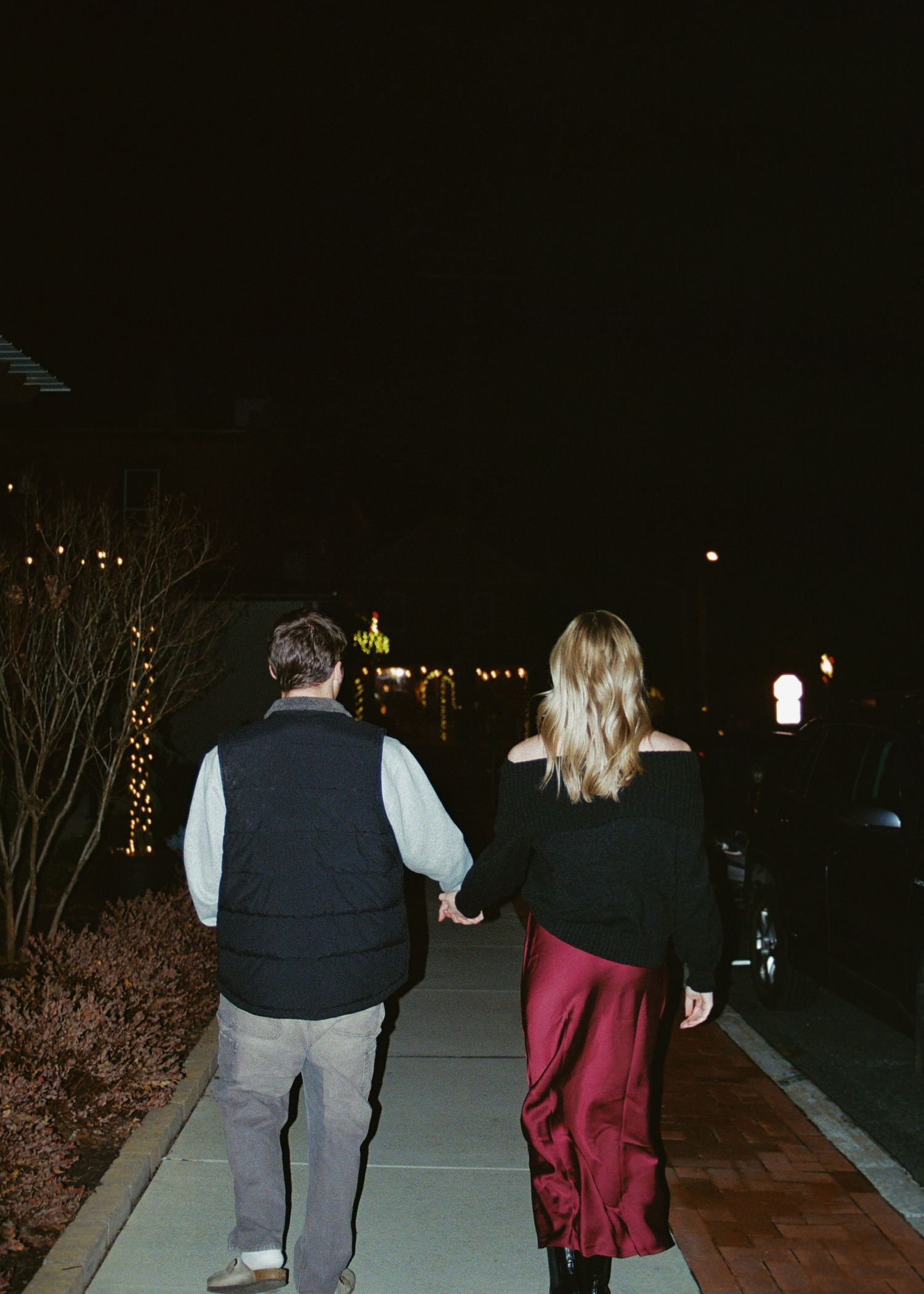 Couple holding hands walking along a lit pathway at night, woman in red satin skirt.