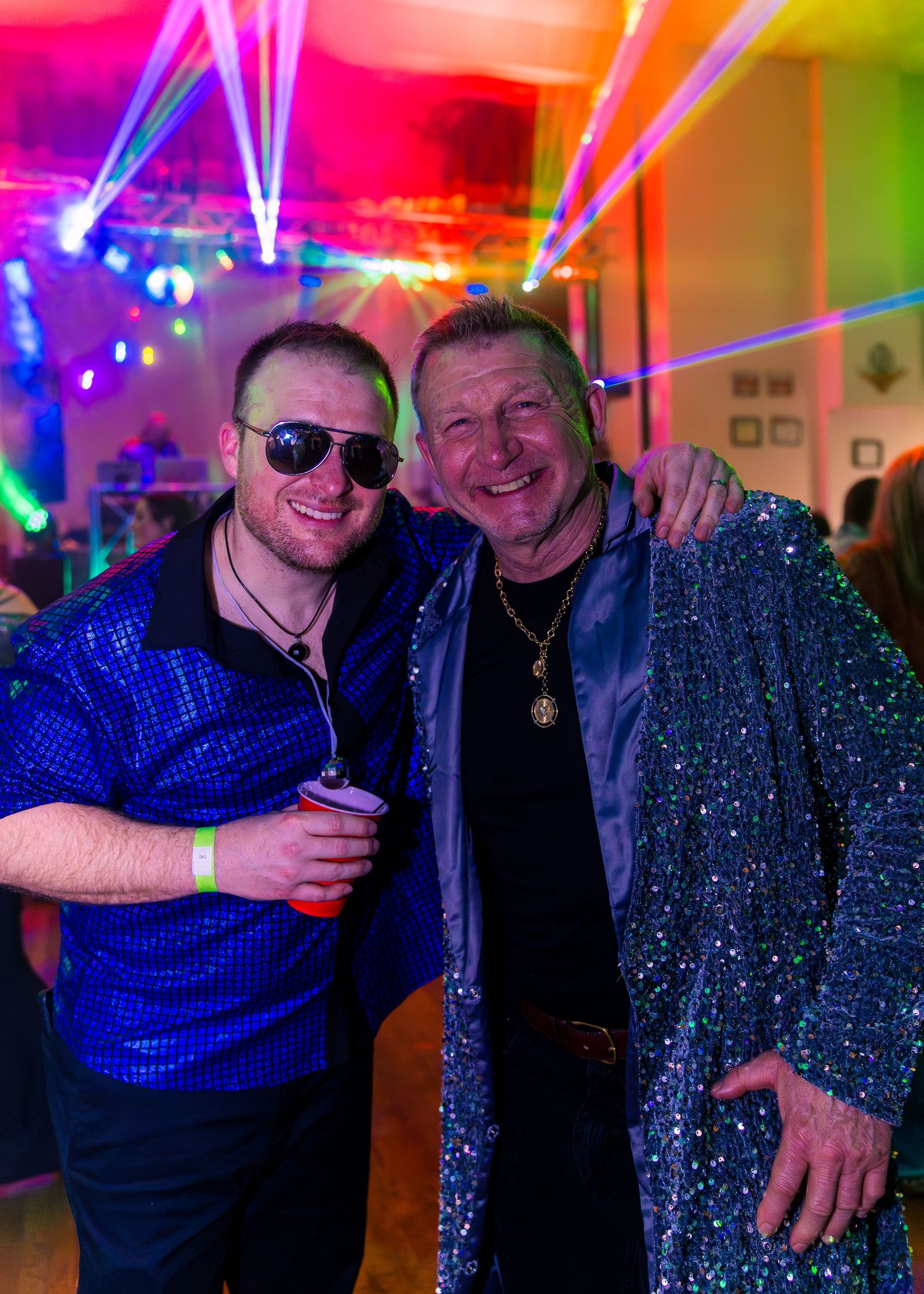 Two smiling men posing together at a colorful disco party with laser lights.
