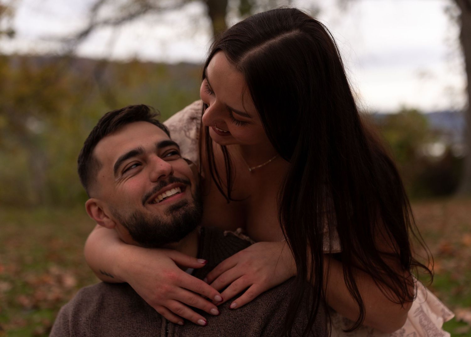 A joyful couple embraces outdoors in autumn, woman hugging man from behind as he smiles warmly.