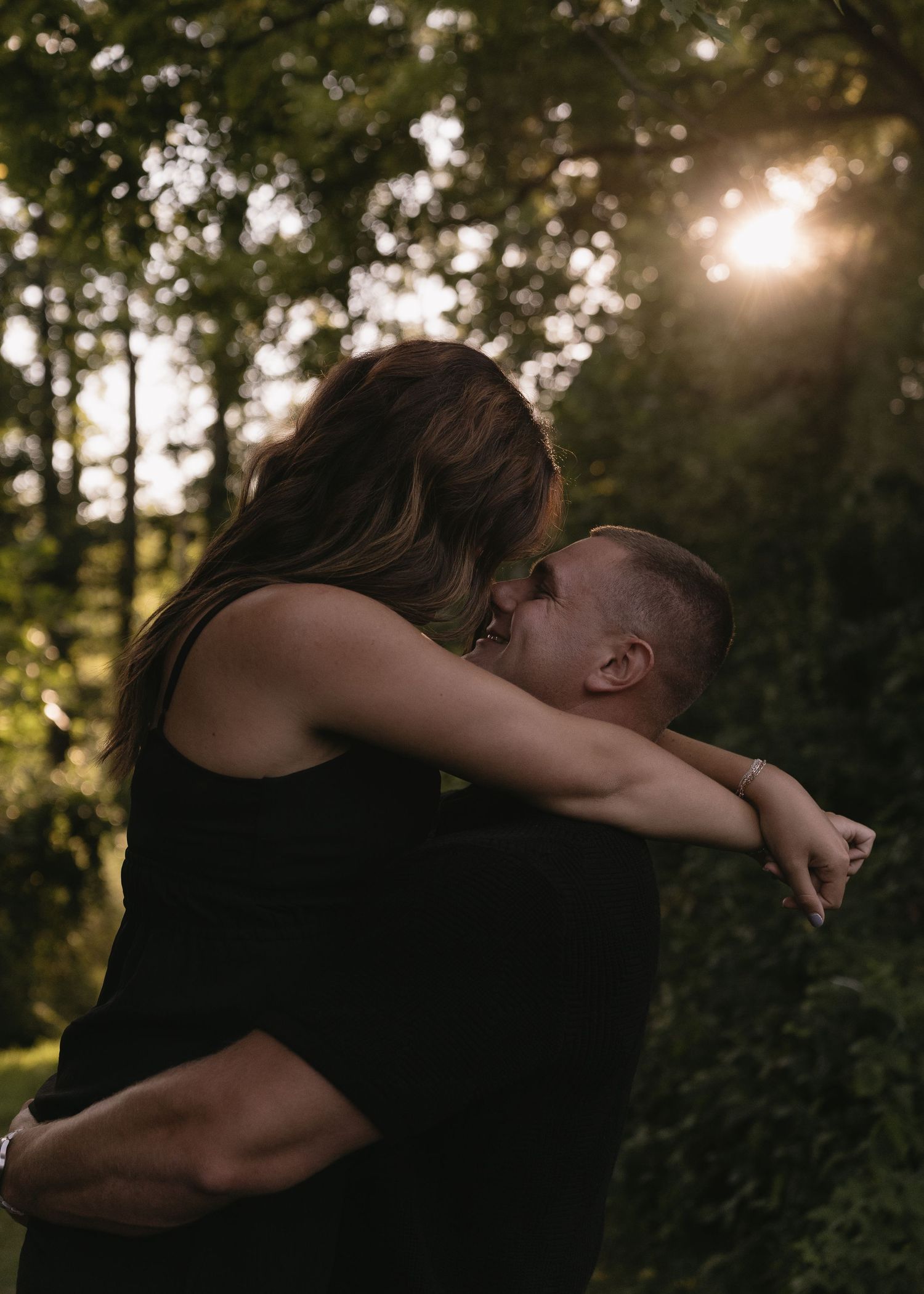 Couple embracing and kissing outdoors at golden hour with sunlight filtering through lush green trees.