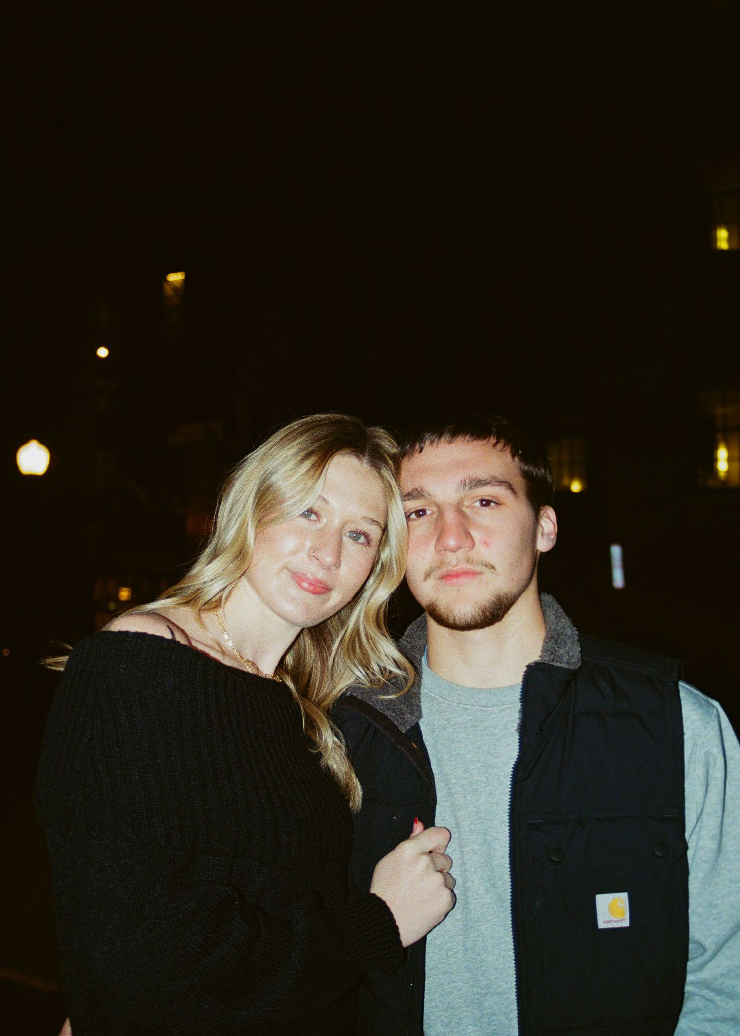 Blonde woman and young man posing together outdoors at night on city street.