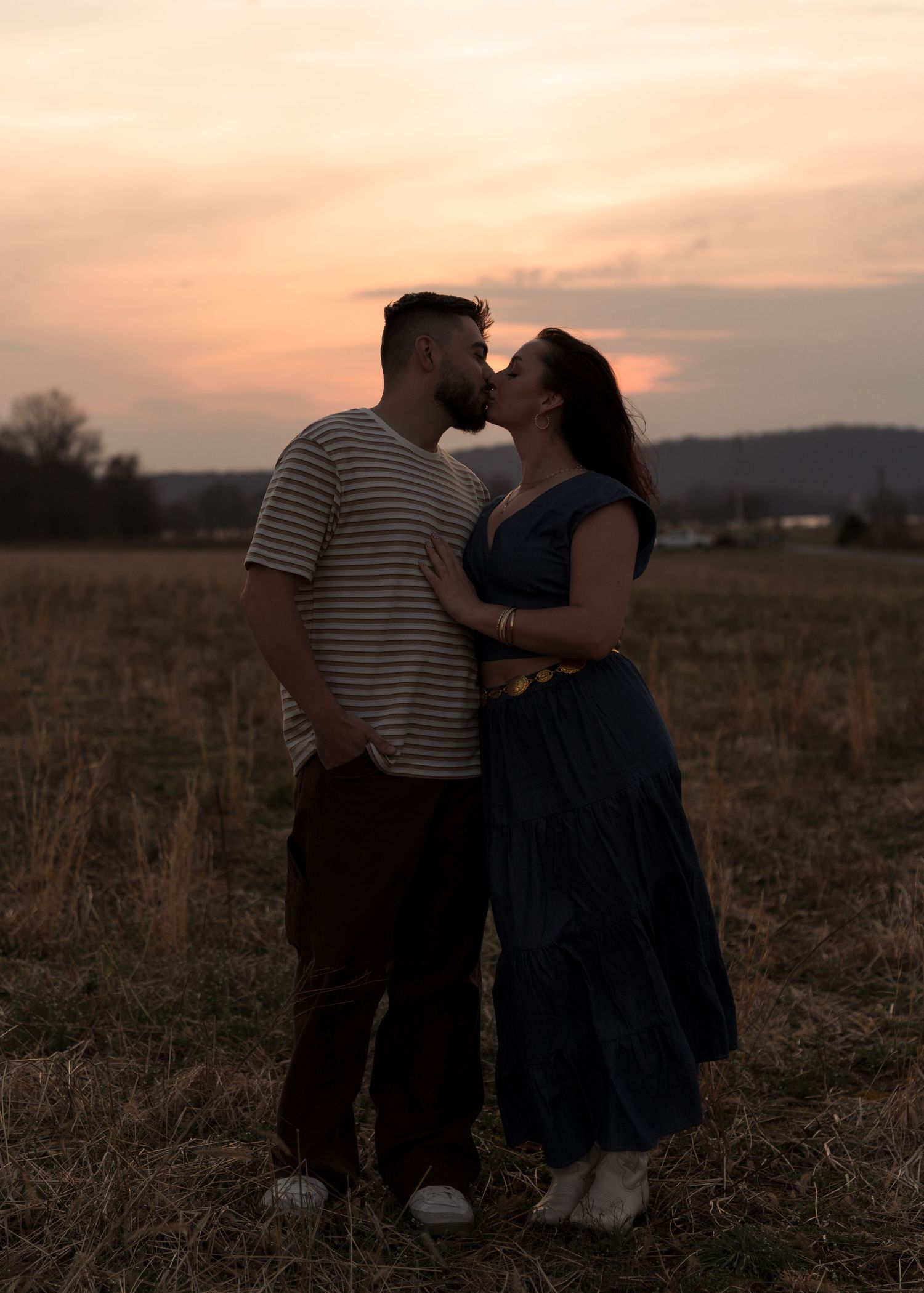 Couple kissing at dusk in open field with dramatic orange sunset sky behind them.