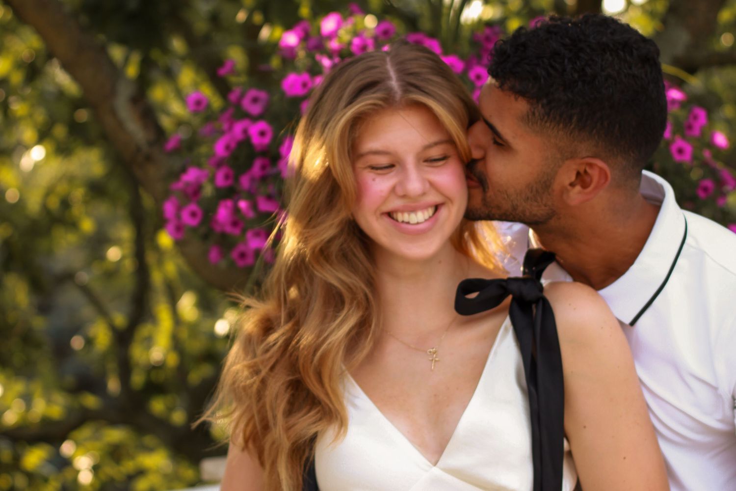 Happy young woman smiling as her partner kisses her cheek near blooming pink flowers.