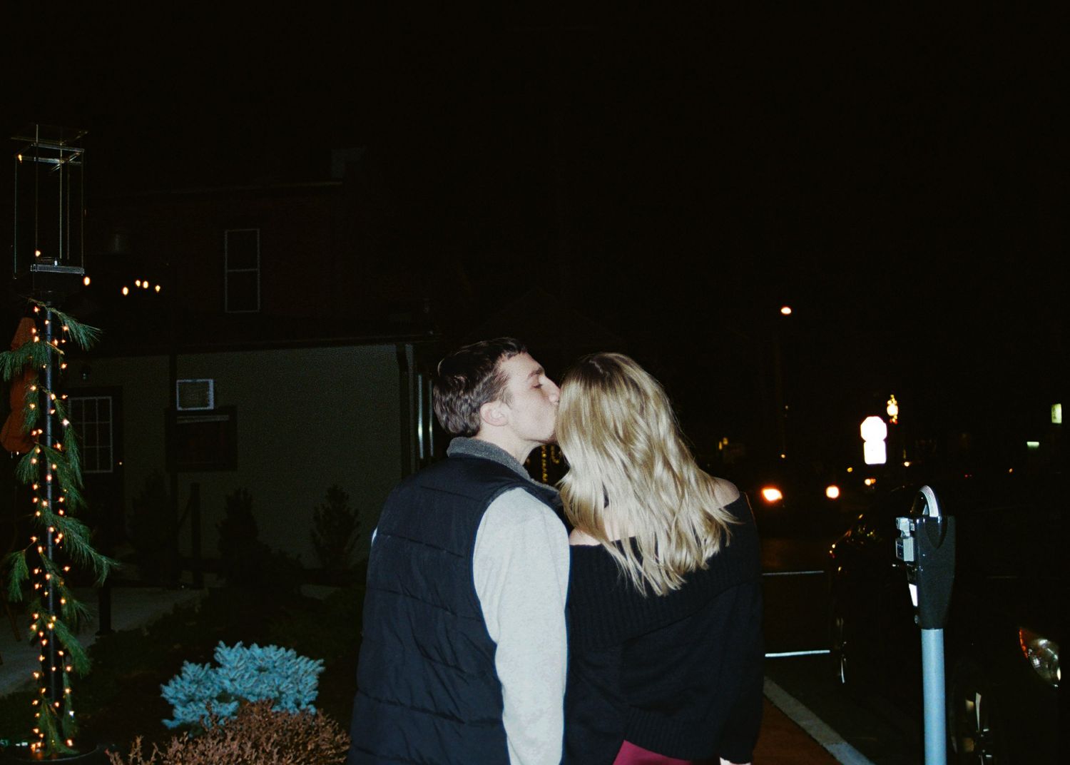 Couple kissing at night near Christmas lights on a city street.