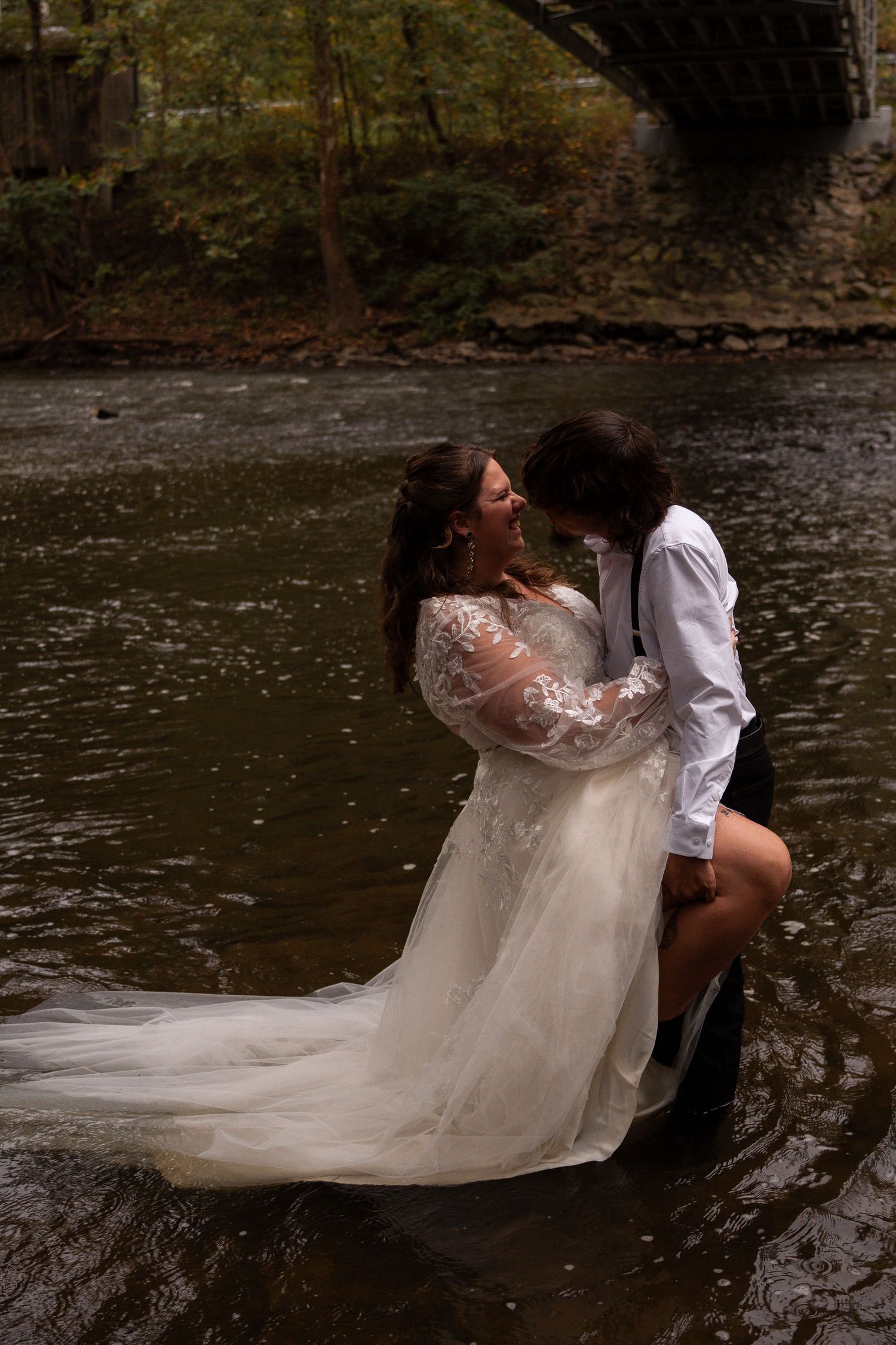 Bride in lace gown and groom embracing in river under stone bridge in autumn.