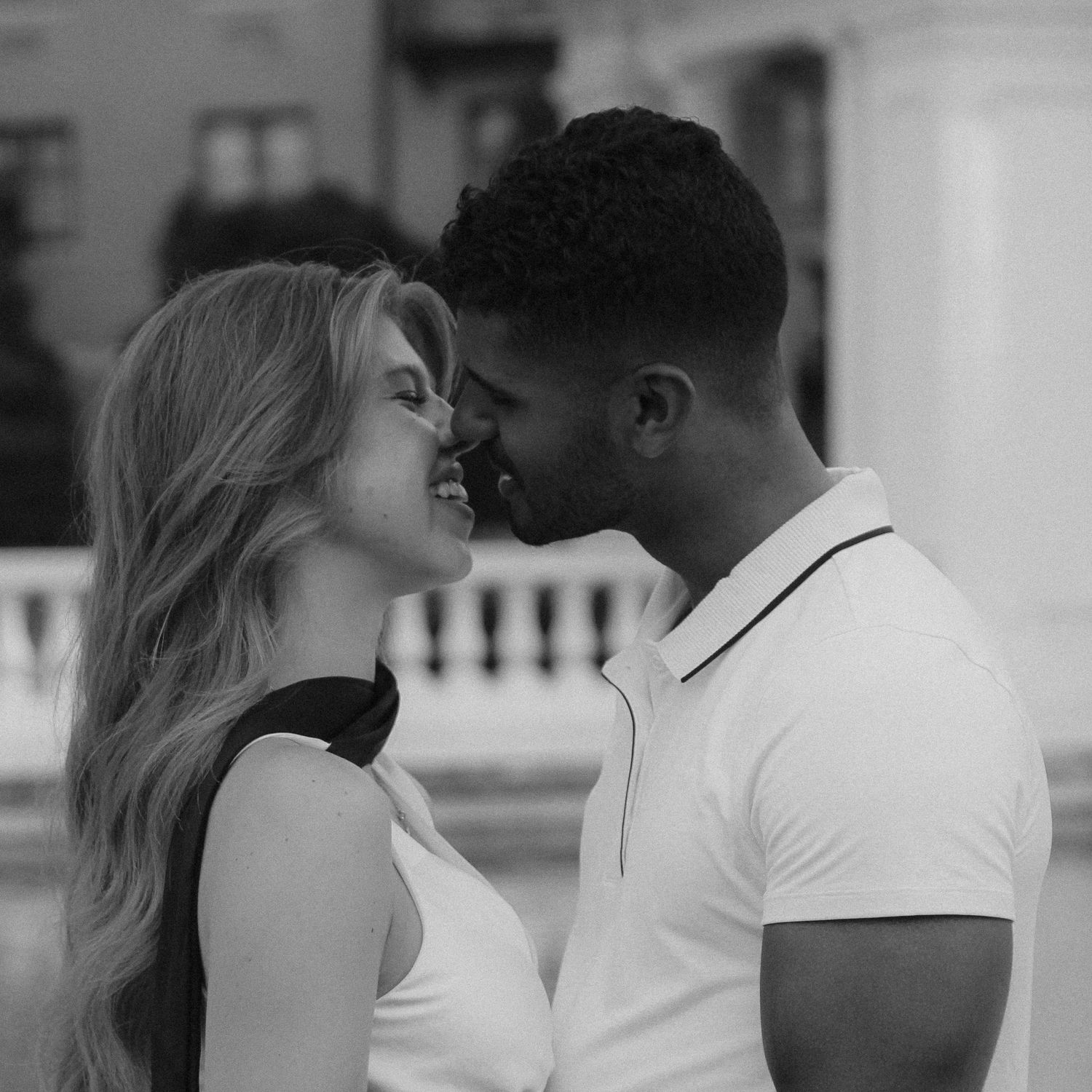 Black and white photo of an interracial couple sharing a romantic moment, nearly kissing outdoors.