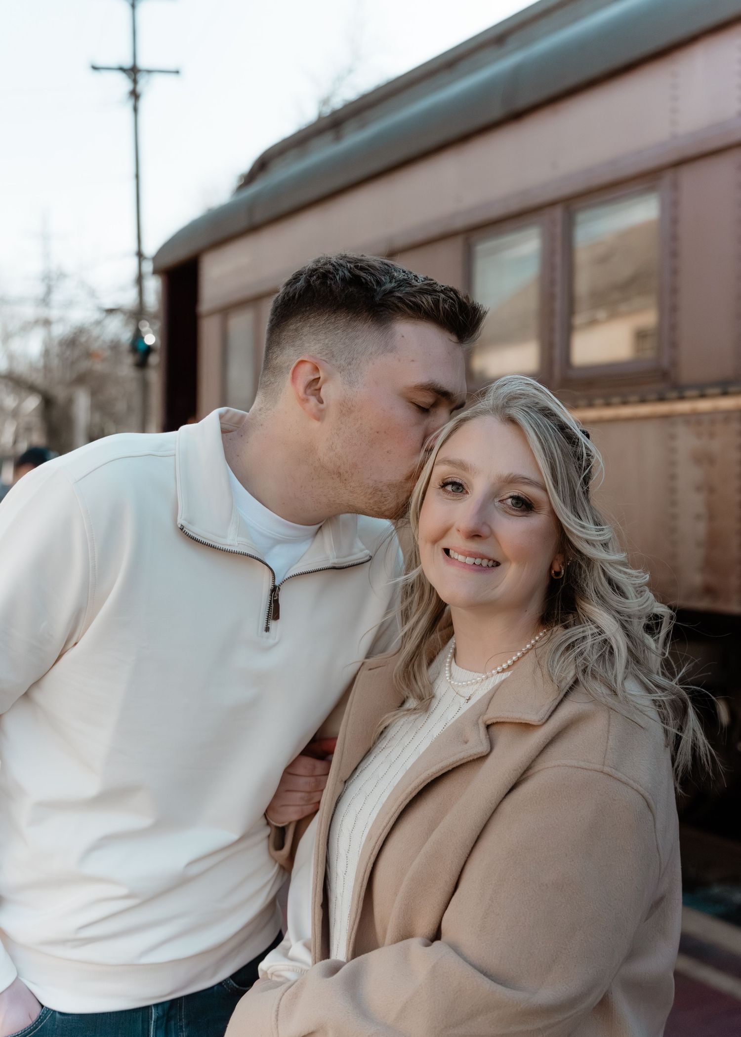 Man kissing smiling woman on the cheek near a vintage train car during engagement photos.