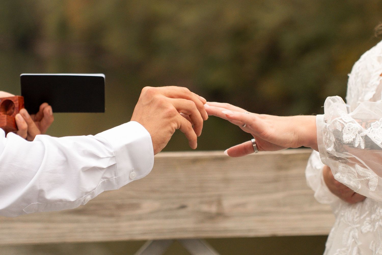 Groom placing wedding ring on bride's finger during outdoor ceremony near water.