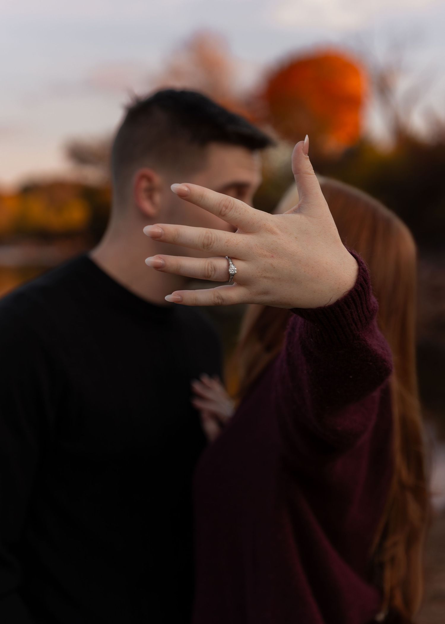 Close-up of woman's hand displaying engagement ring with couple embracing in fall background.