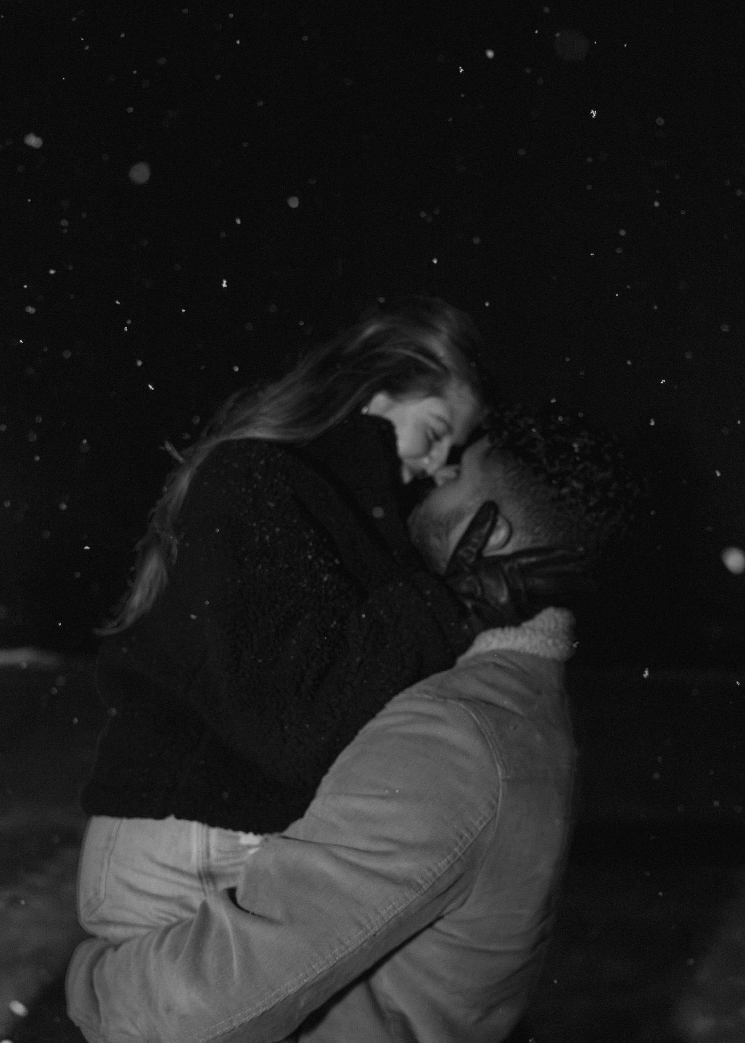 Black and white romantic couple embracing in falling snow at night, sharing an intimate moment outdoors.