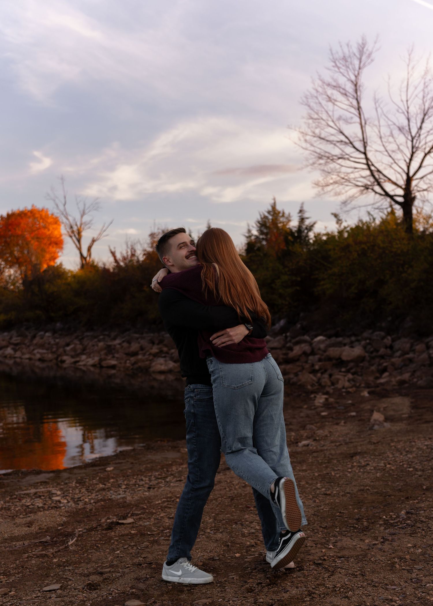 Couple embracing and laughing together near a lake during golden hour in autumn.
