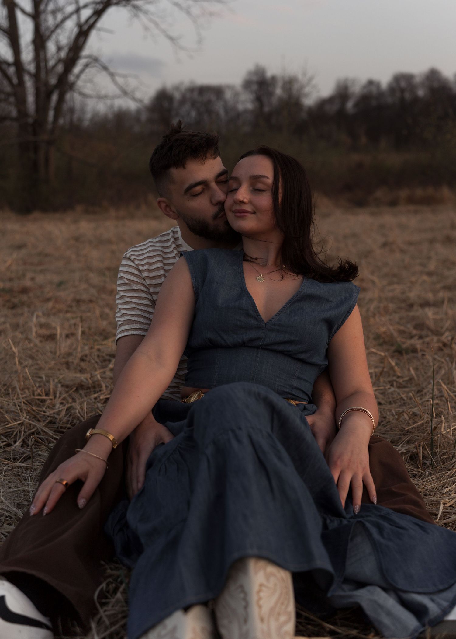 Couple sitting in a dry field at dusk, man kissing woman's cheek as she smiles.