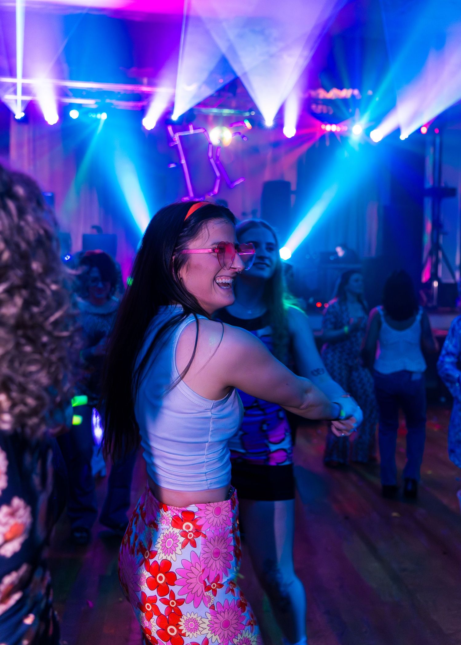 Young woman dancing joyfully in floral skirt and pink sunglasses at a vibrant nightclub.