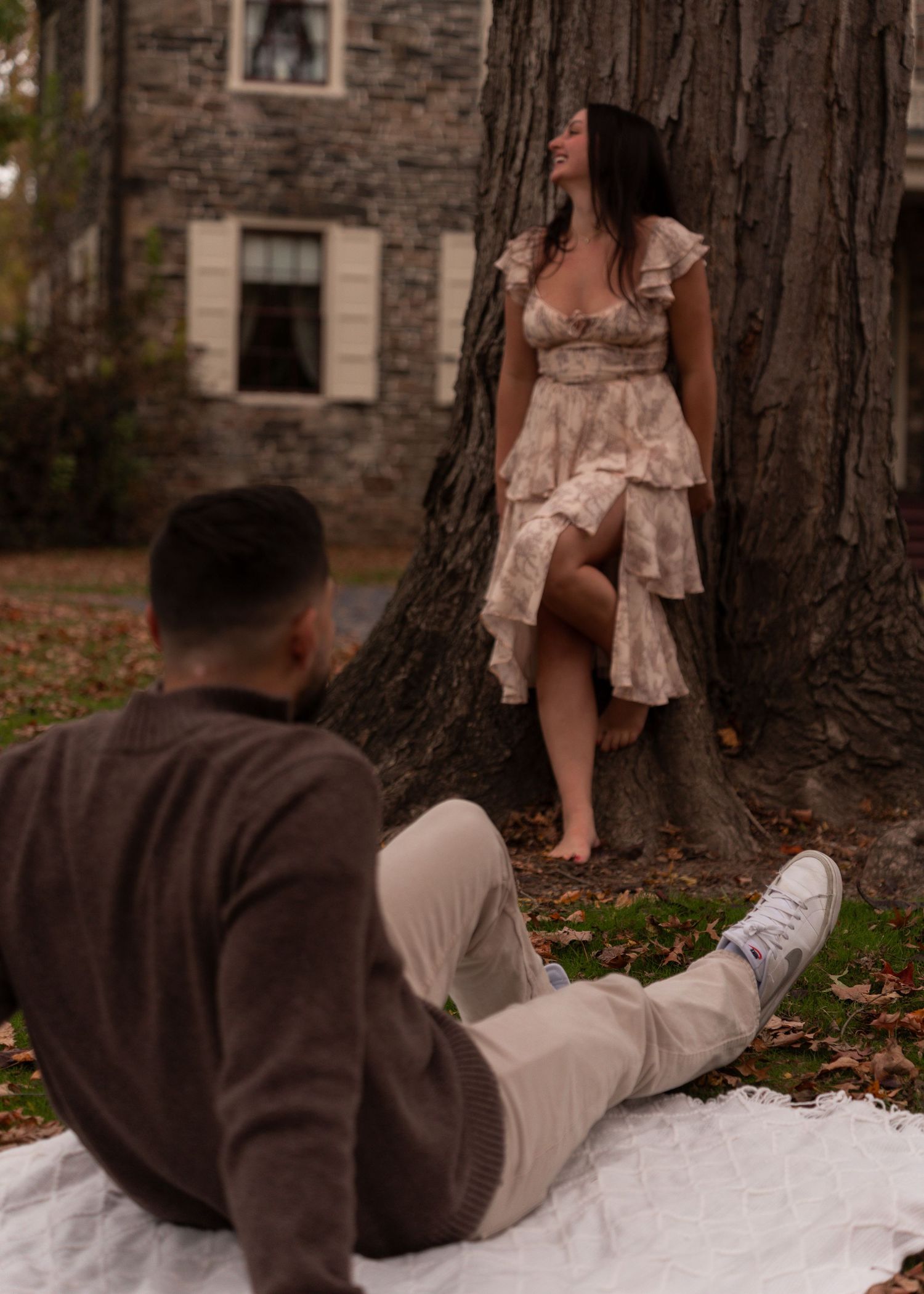 A woman in a floral dress leans against a tree while her partner sits on a blanket.