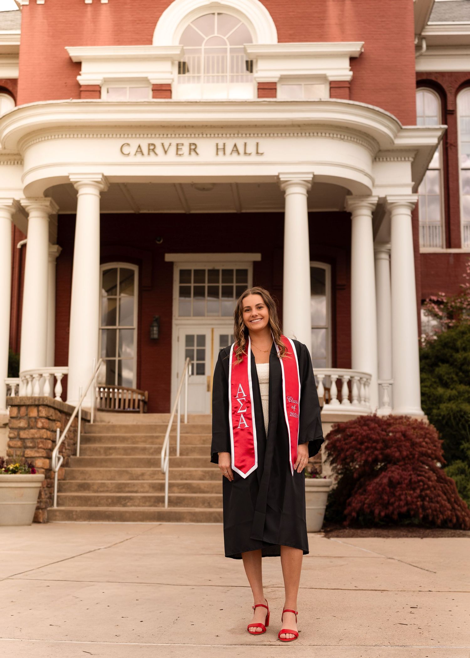 Smiling graduate in cap and gown with red sash standing before Carver Hall.