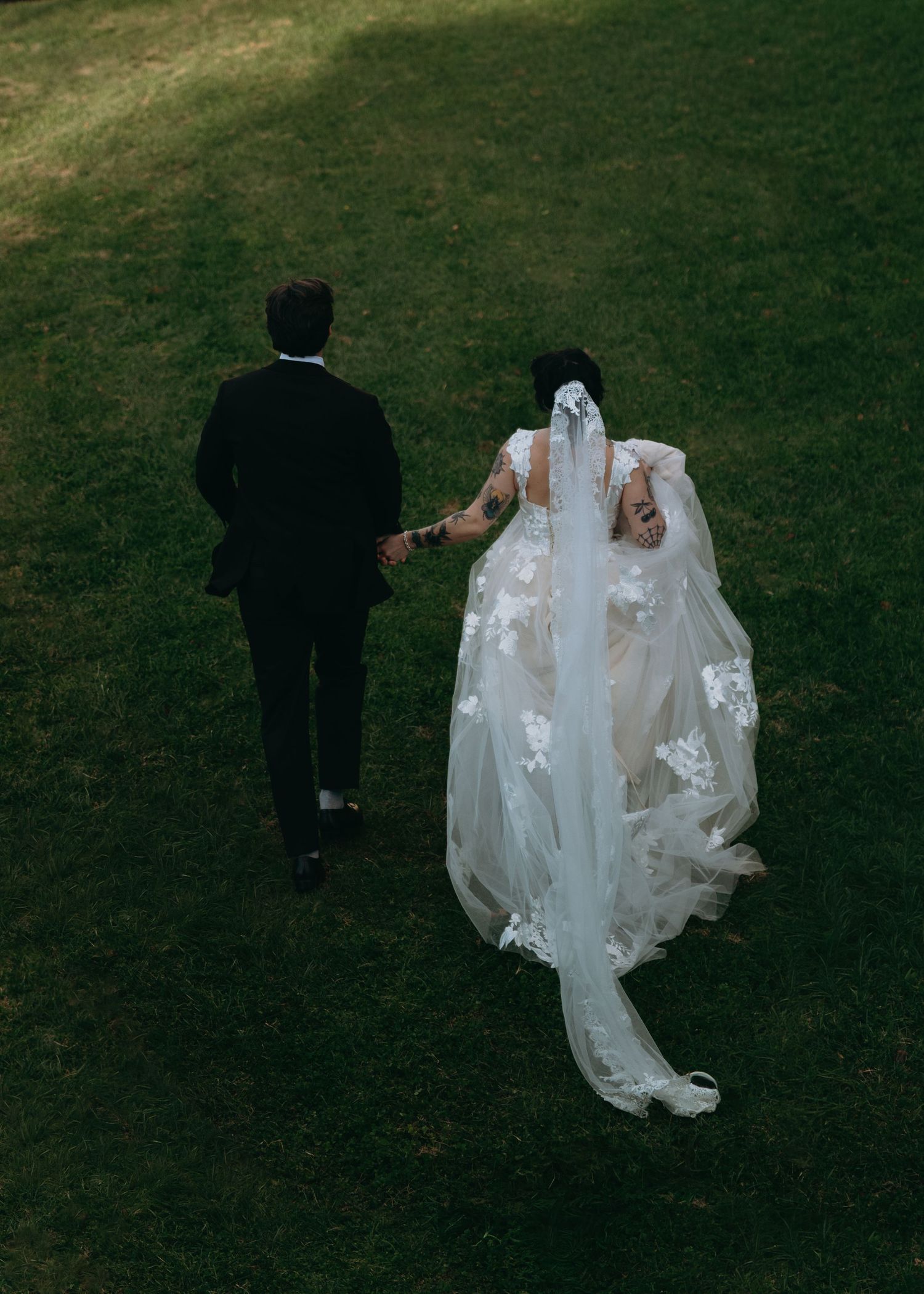 Aerial view of bride and groom holding hands walking across green grass, bride wearing lace cathedral veil.