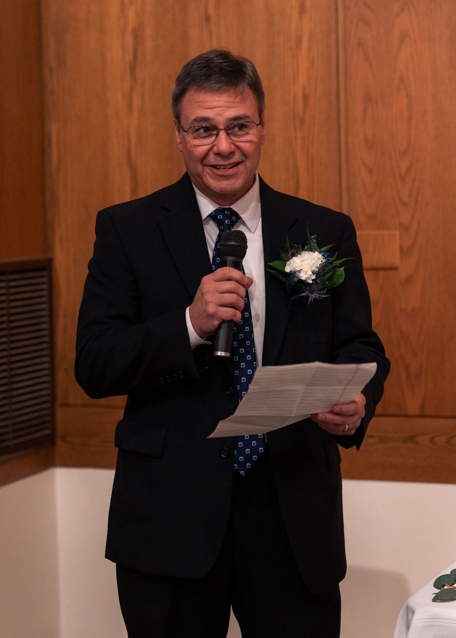 Smiling man in black suit giving a wedding speech into microphone while holding notes at reception.