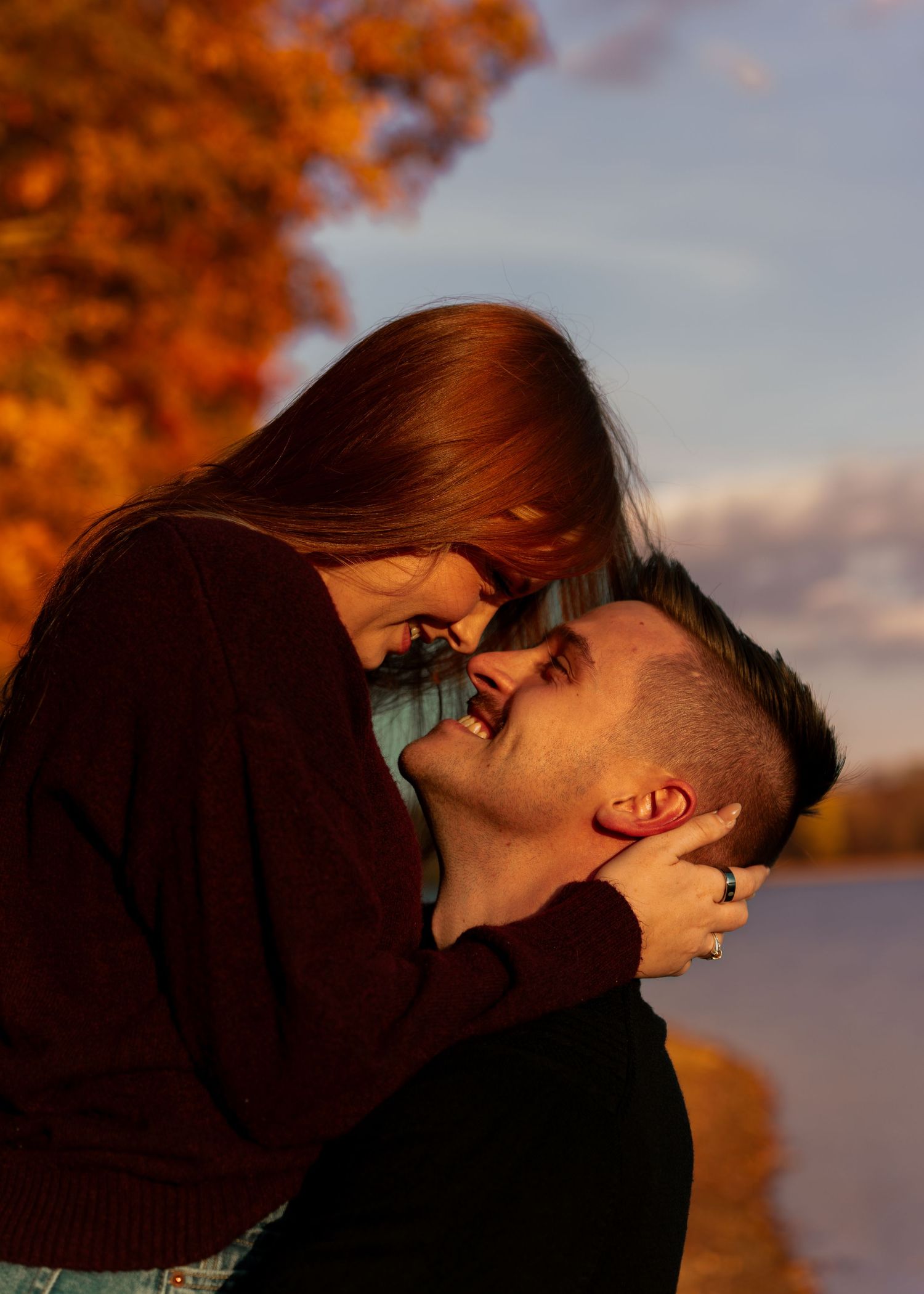 Woman tenderly holds mans face while leaning in close, framed by vibrant autumn foliage at sunset.