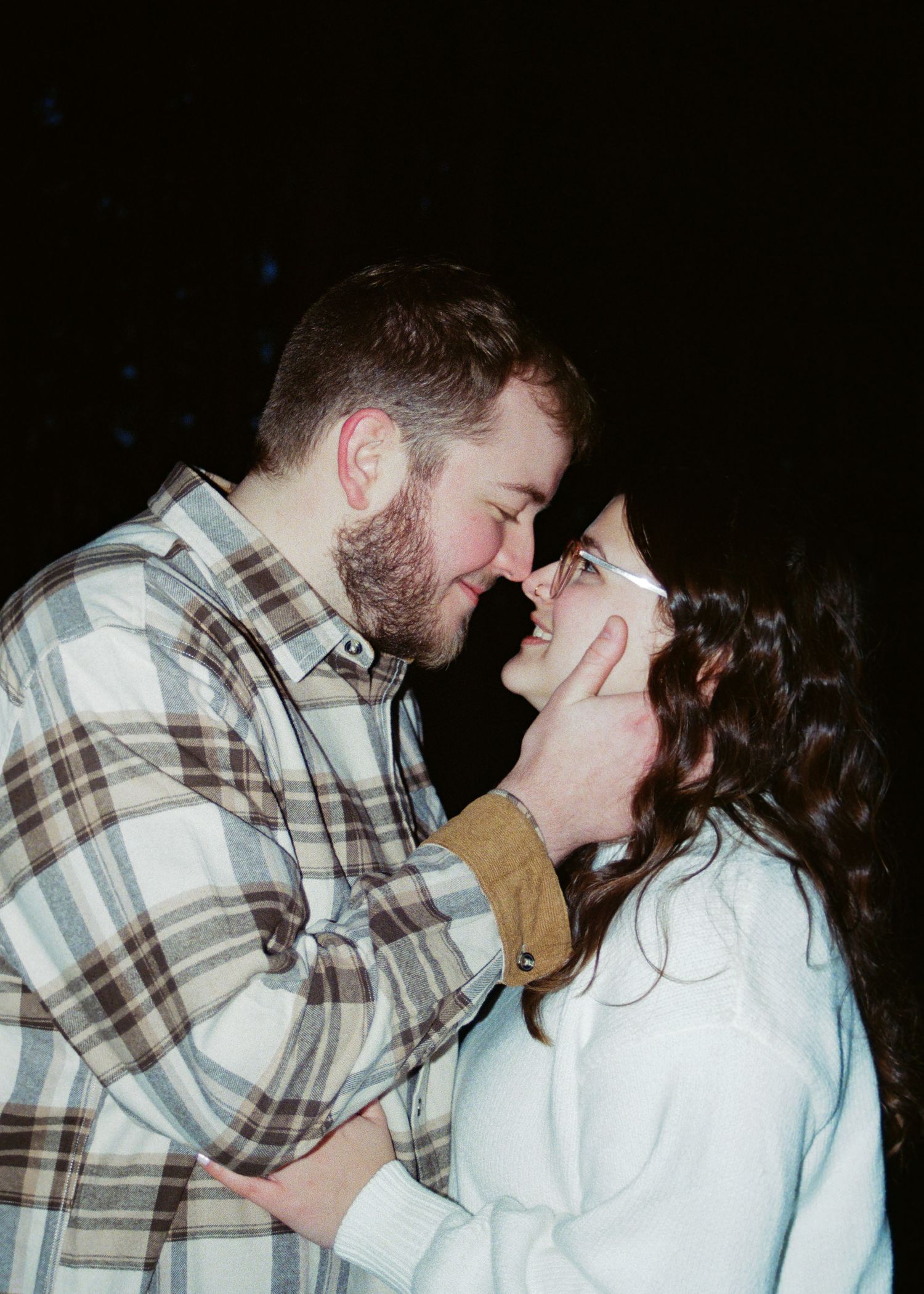 Couple sharing an intimate moment outdoors at night, man with beard and woman wearing glasses smiling.