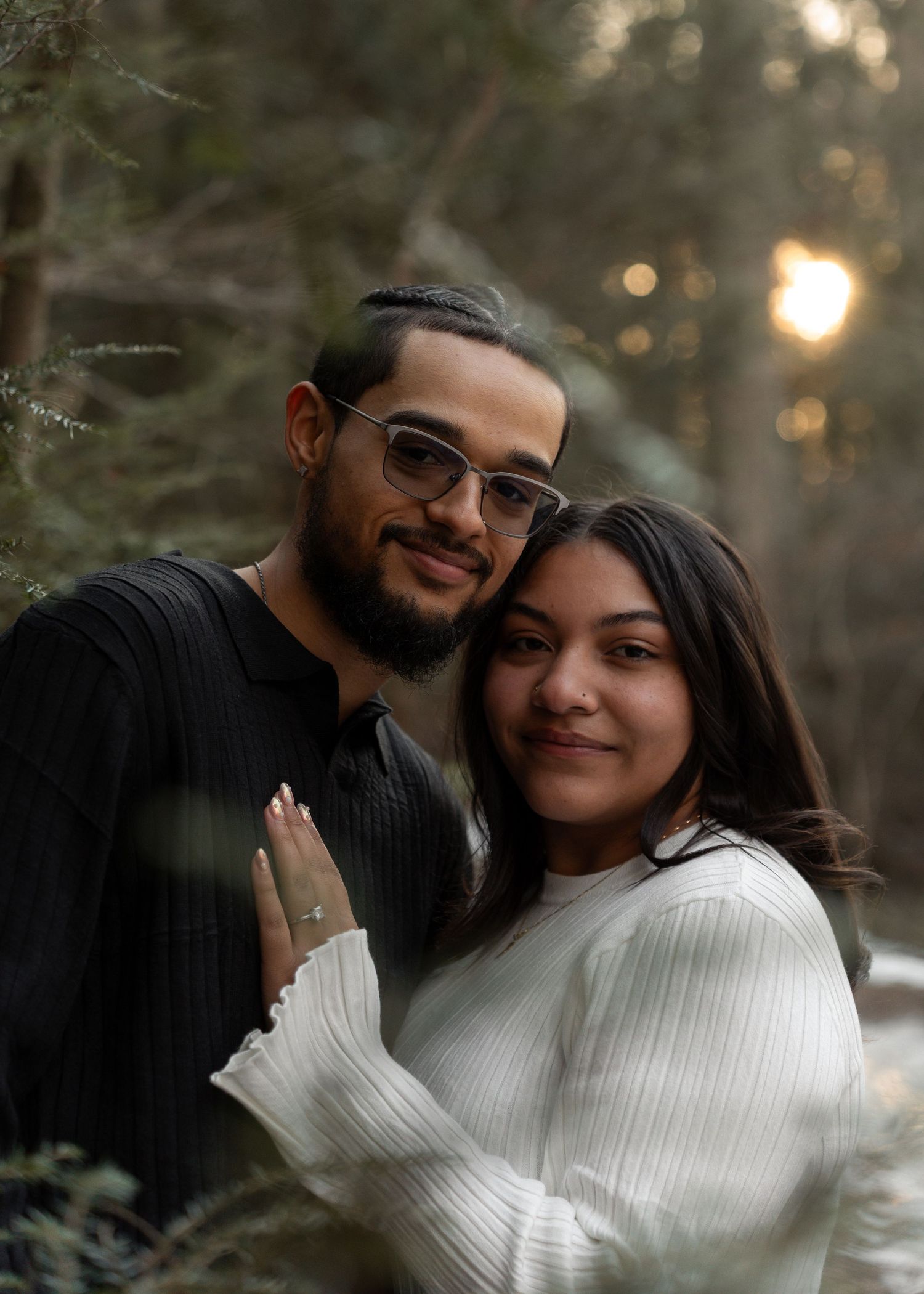 Smiling engaged couple posing together in forest at sunset, man in black shirt and woman in white sweater.