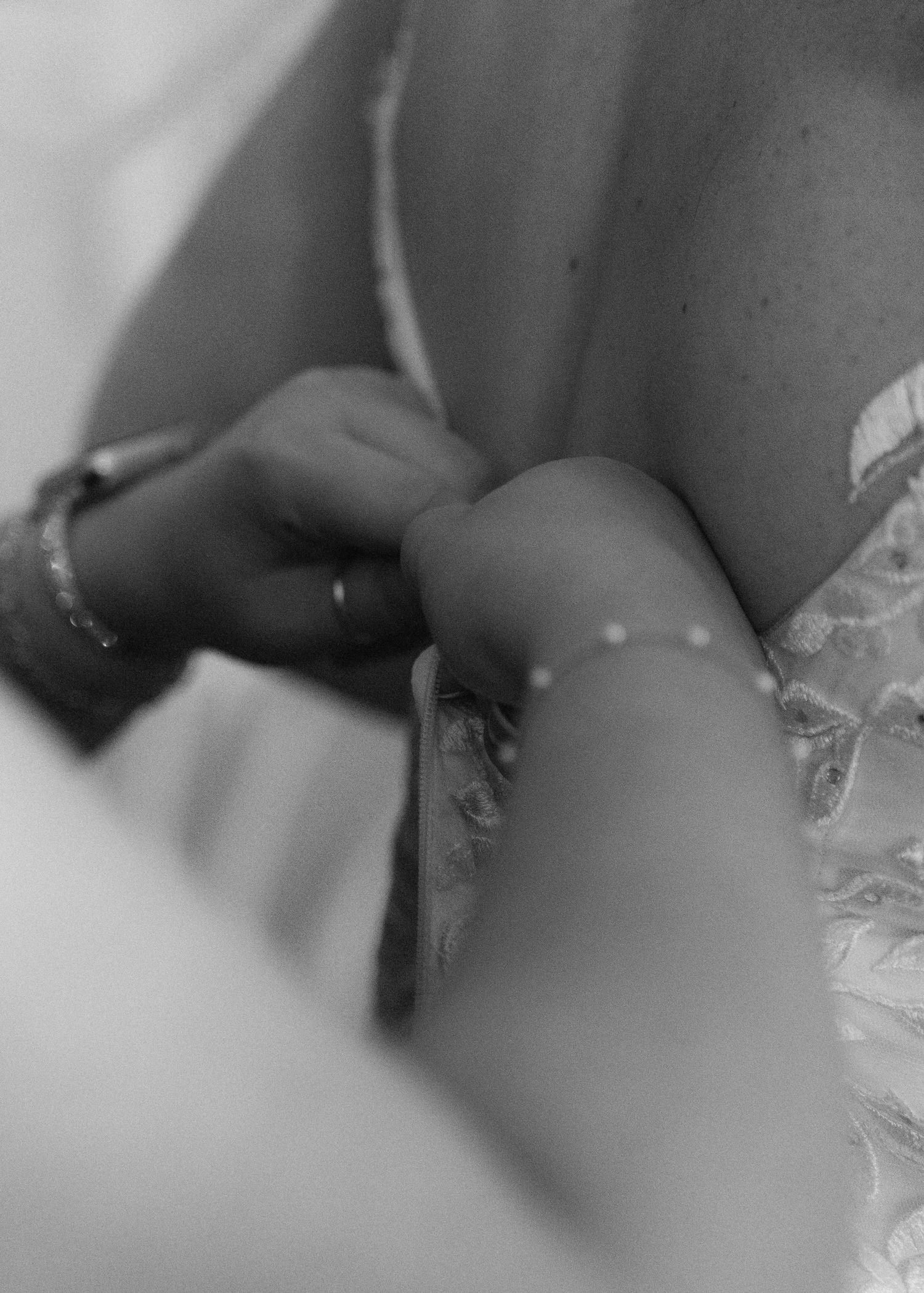 Black and white detail of hands fastening pearl buttons on back of lace wedding dress.