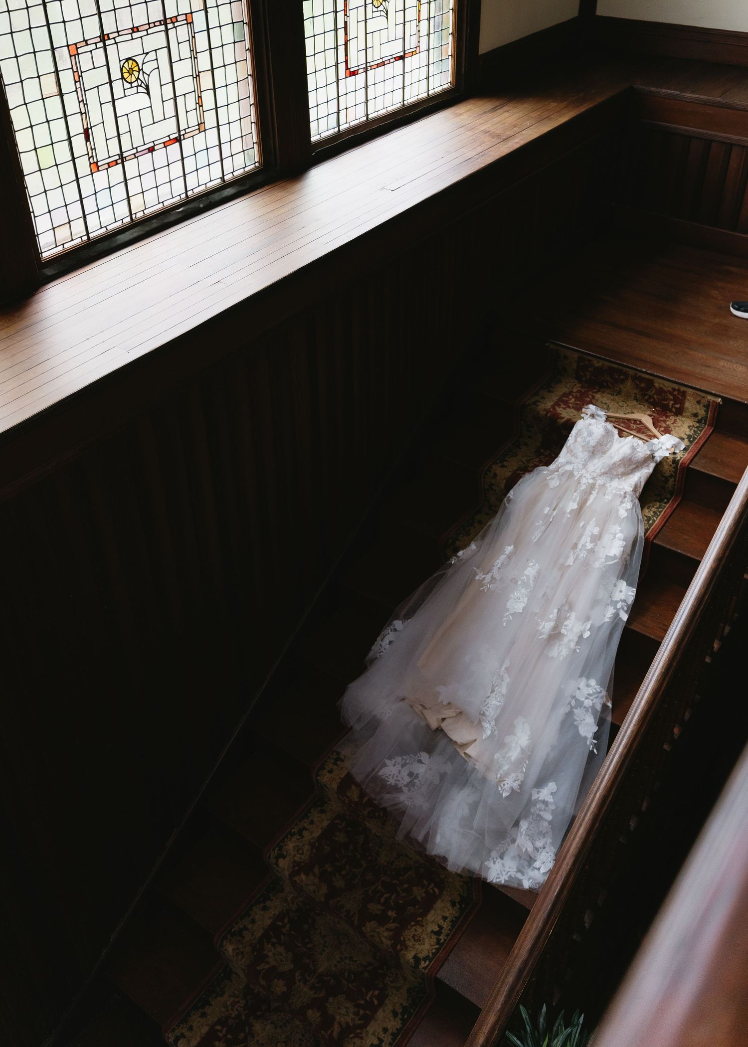 Elegant lace wedding dress hanging on wooden staircase near stained glass windows in vintage venue.