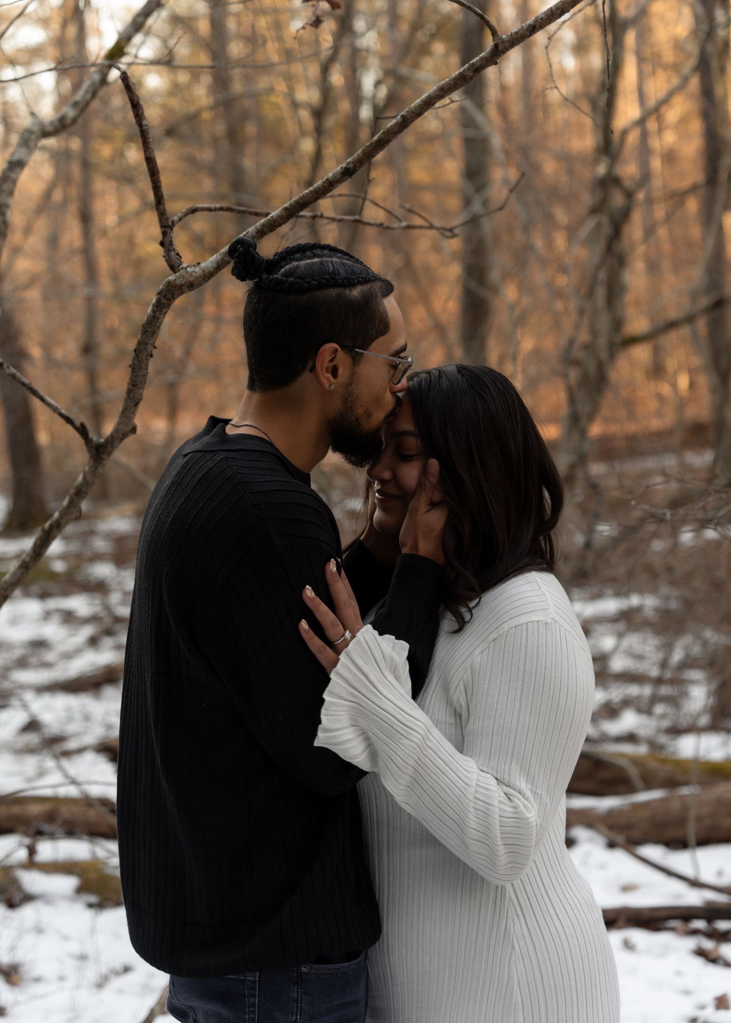 Romantic couple nearly kissing in snowy winter woods, man in black and woman in white ribbed sweater.