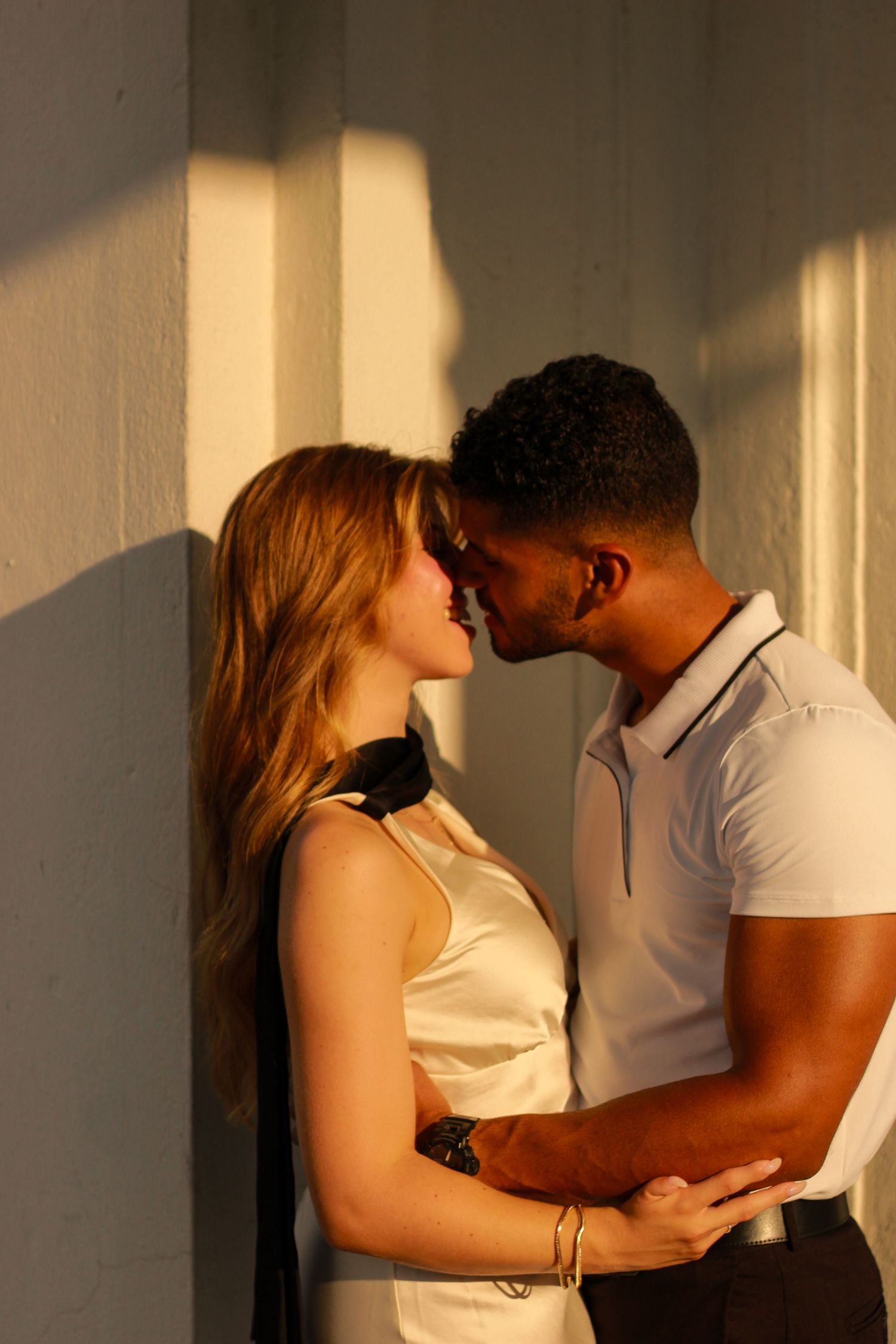 Interracial couple leans in for a kiss against a sunlit wall, bathed in warm golden light.