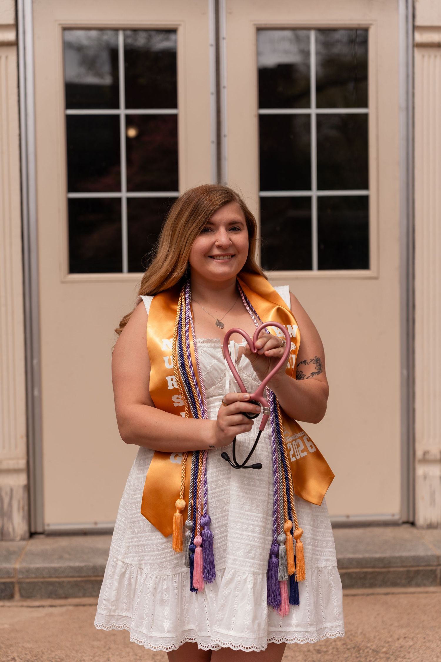 Smiling nursing graduate in white dress holding stethoscope with gold and purple honor cords.