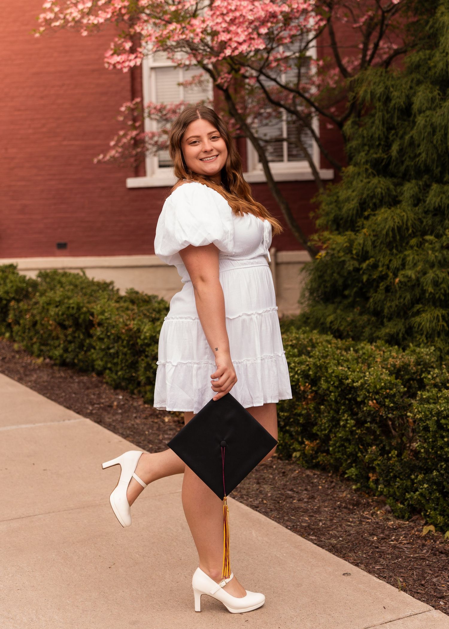 Smiling graduate in white dress holding a cap with gold tassel near blooming trees.