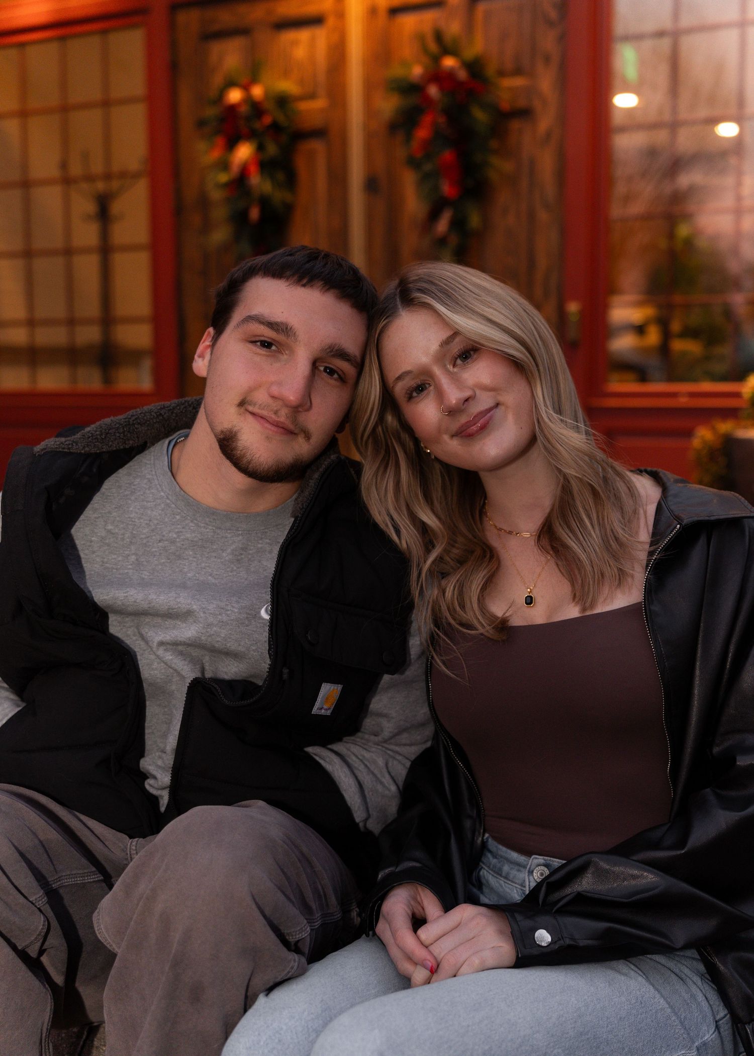 Smiling couple leaning together outdoors near festive holiday wreaths on a red door.