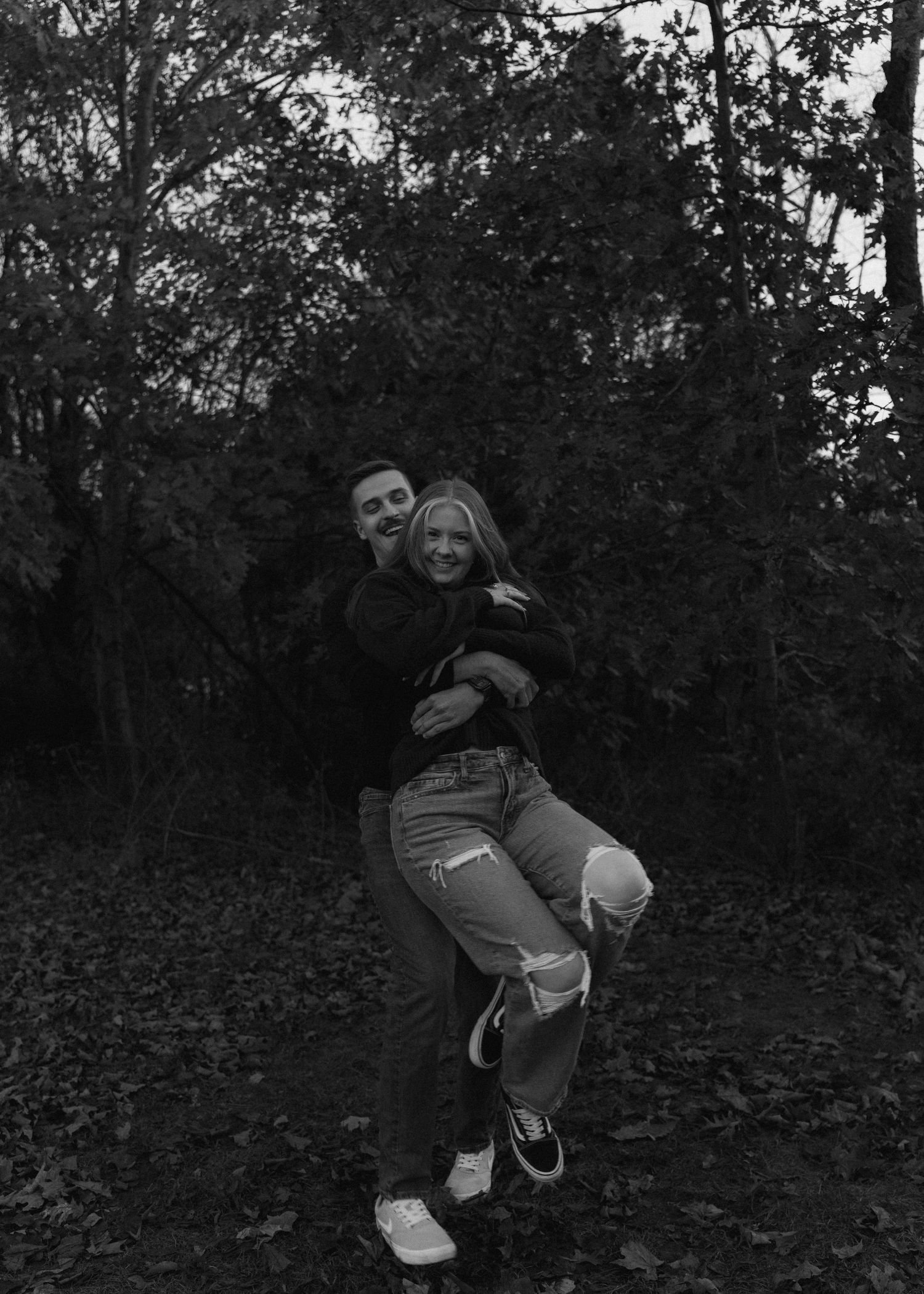 Black and white photo of happy couple embracing and laughing together in a wooded autumn setting.