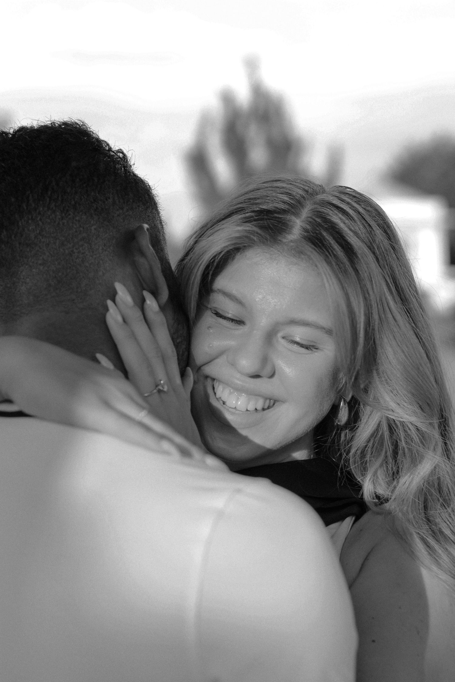 Black and white photo of a joyful woman smiling while embracing her partner outdoors.
