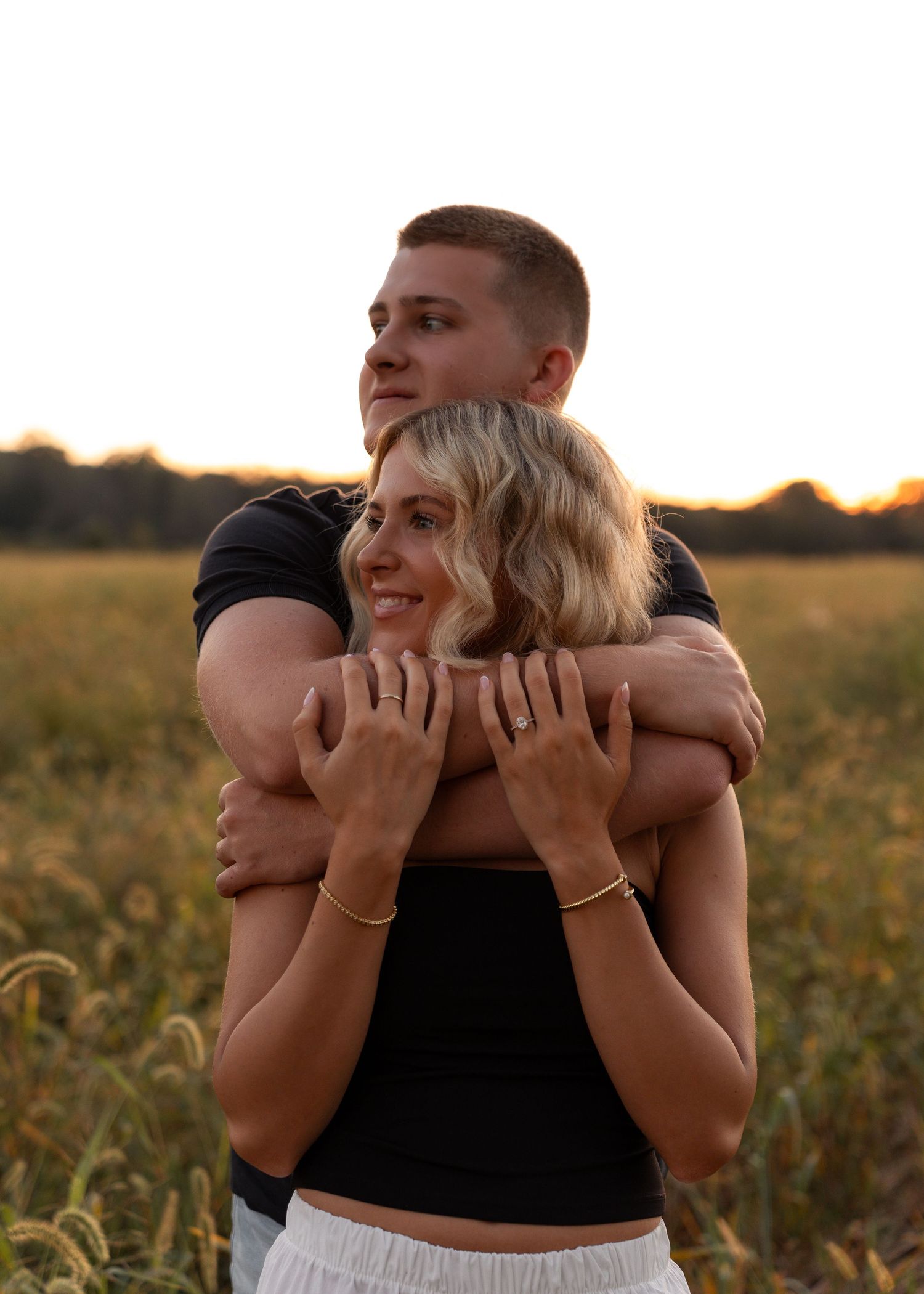 A couple embraces warmly in a golden wheat field at sunset, man wrapping arms around smiling woman.