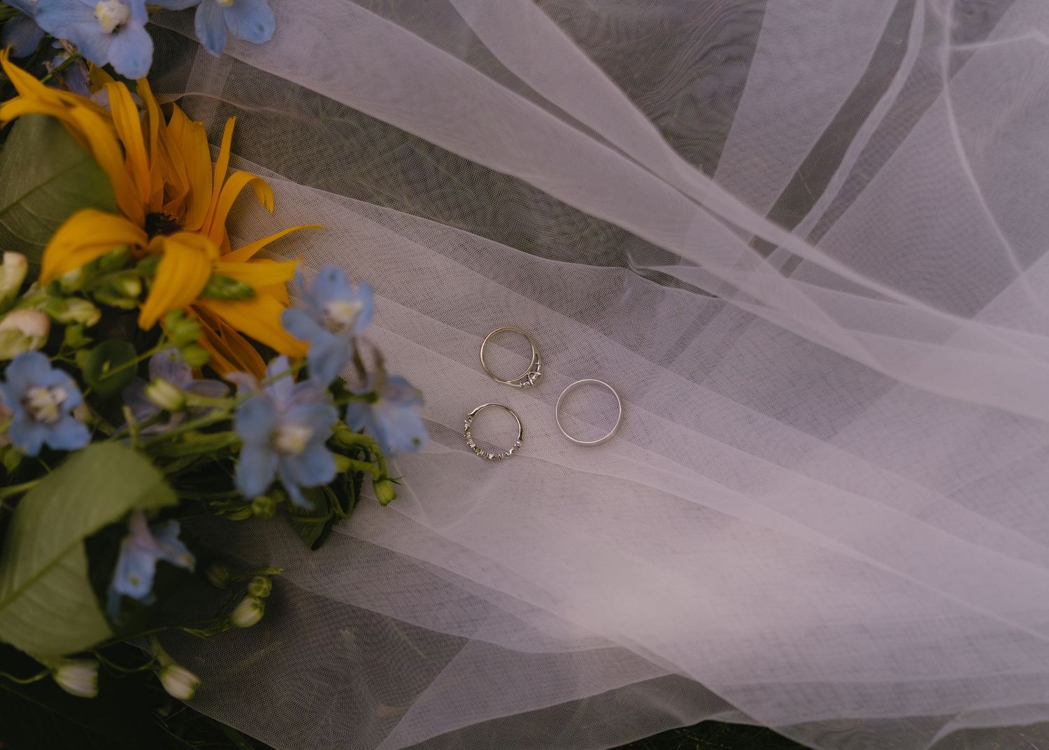 Wedding rings resting on white tulle veil beside a bouquet of yellow sunflowers and blue forget-me-nots.